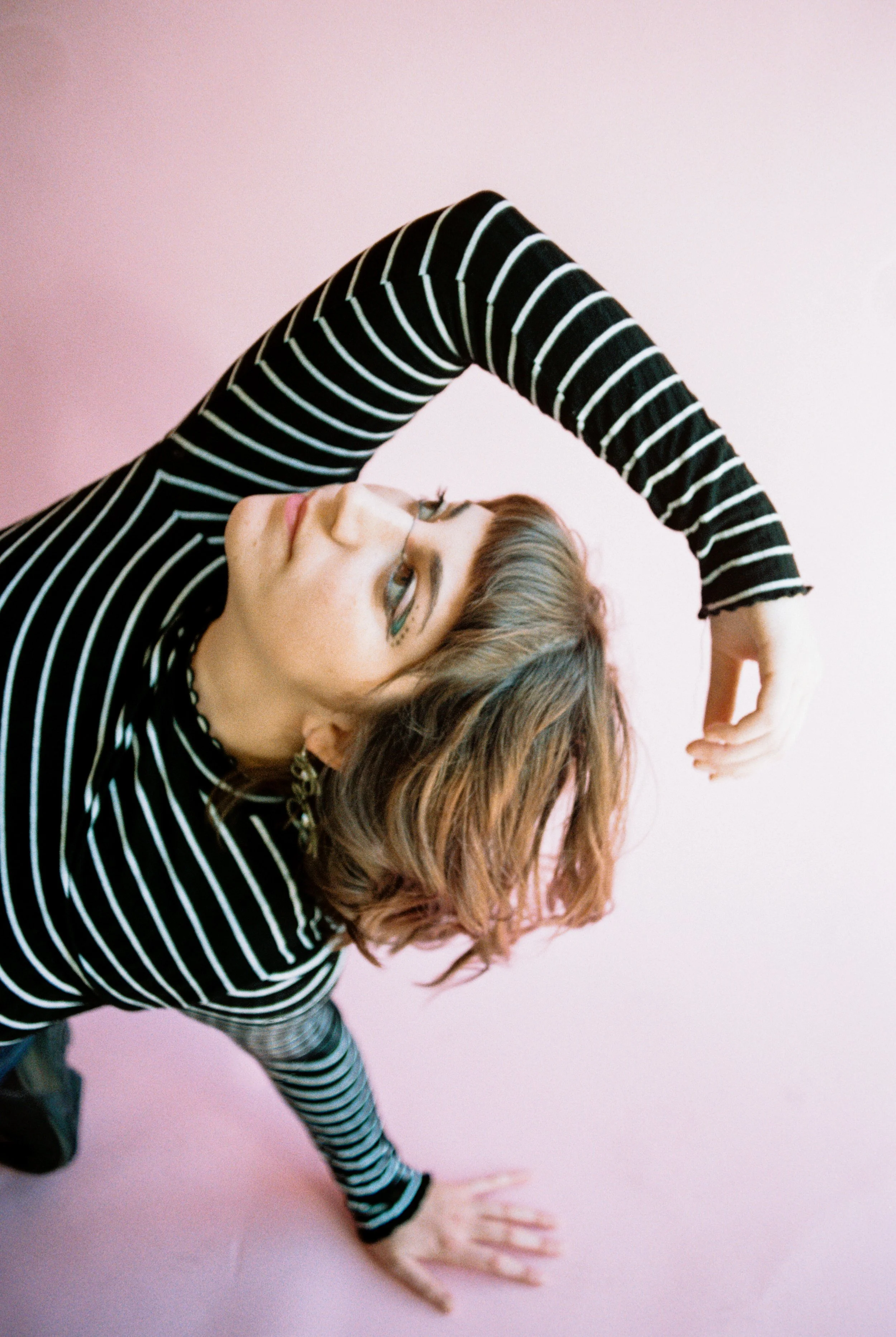 A person with brown, wavy hair doing a yoga pose against a pink background, wearing a black and white striped long-sleeve shirt.