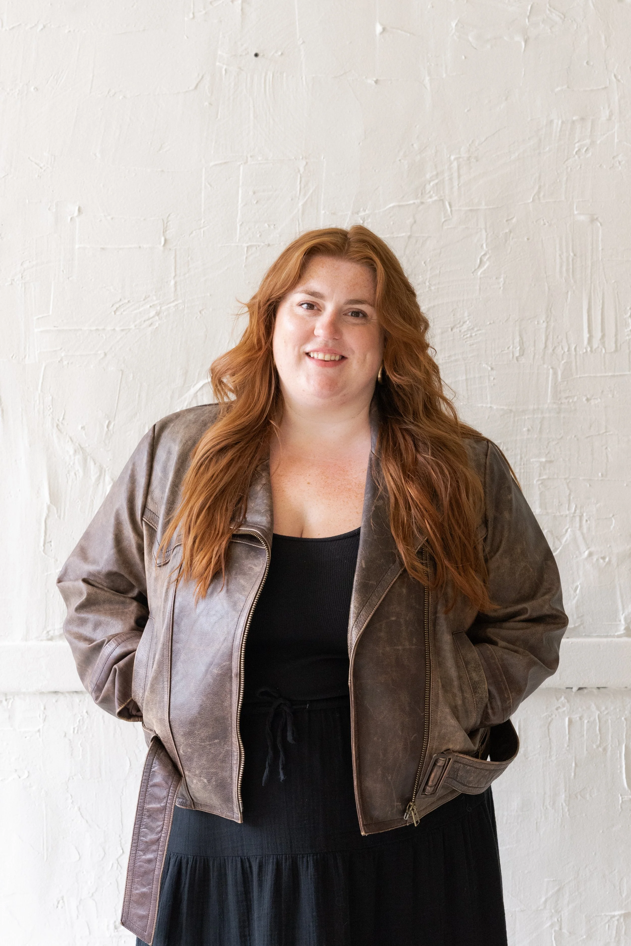 A woman with long red hair smiling, wearing a brown leather jacket over a black top and a black skirt, standing in front of a white brick wall.