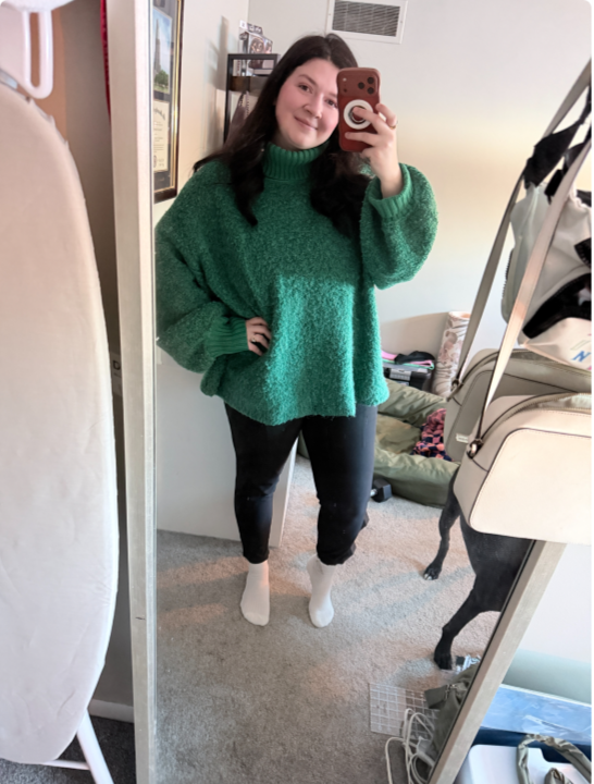 Young woman taking a mirror selfie in a room, wearing a green oversized sweater, black ripped jeans, and white socks, smiling.