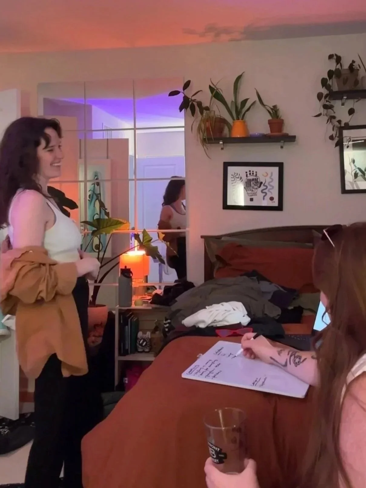 A woman stands talking in a room with a bed and a person sitting at a table writing on a notepad. The room is decorated with plants, framed pictures, and colorful lighting.