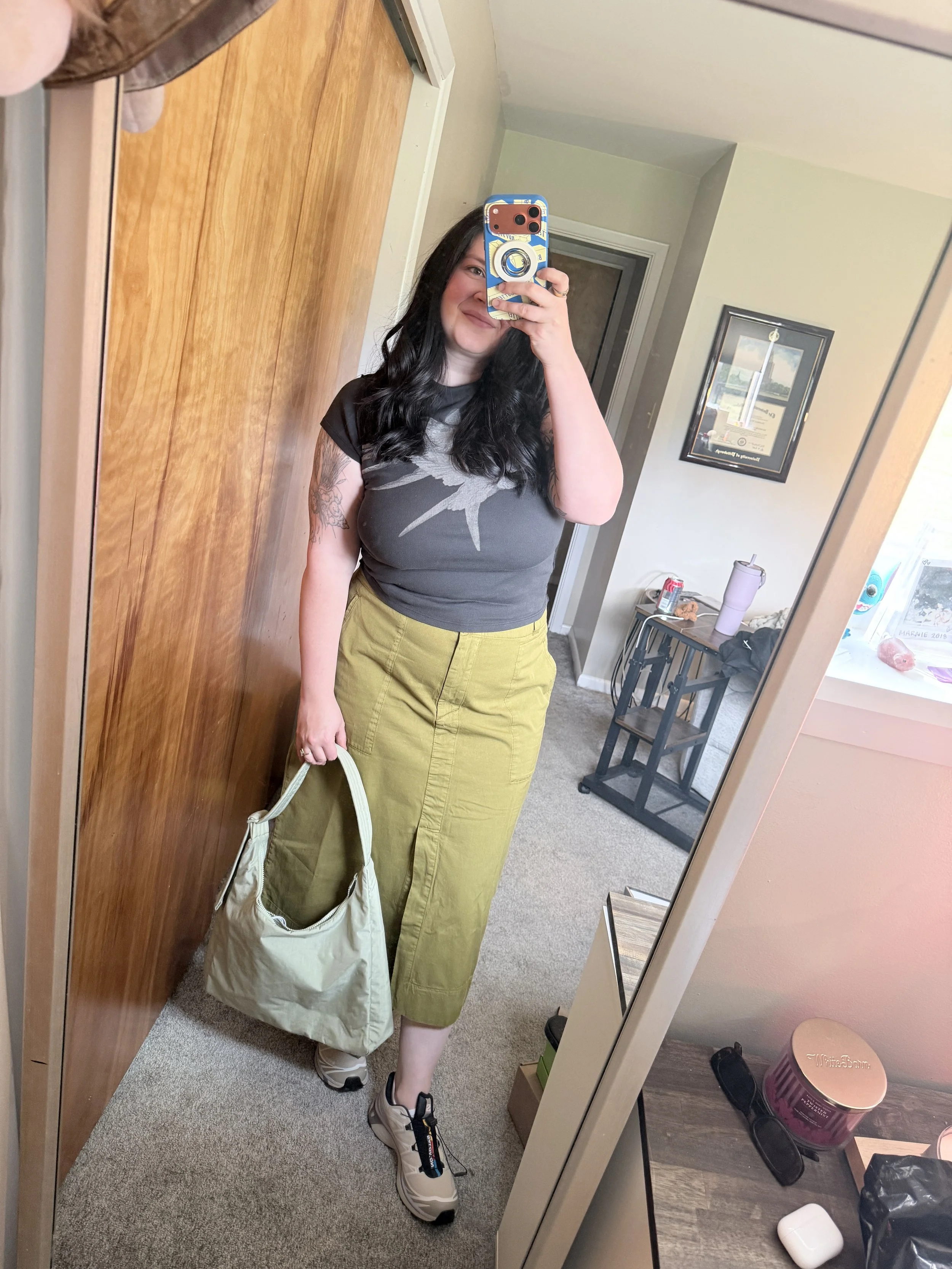 A woman taking a mirror selfie in a room. She has black wavy hair and is wearing a black and gray t-shirt with a bird graphic, olive green pants, and beige sneakers. She is holding a beige bag and smiling. The room has a wooden door, a small table with a water bottle, a can of soda, and various items, and is decorated with framed pictures on the walls.