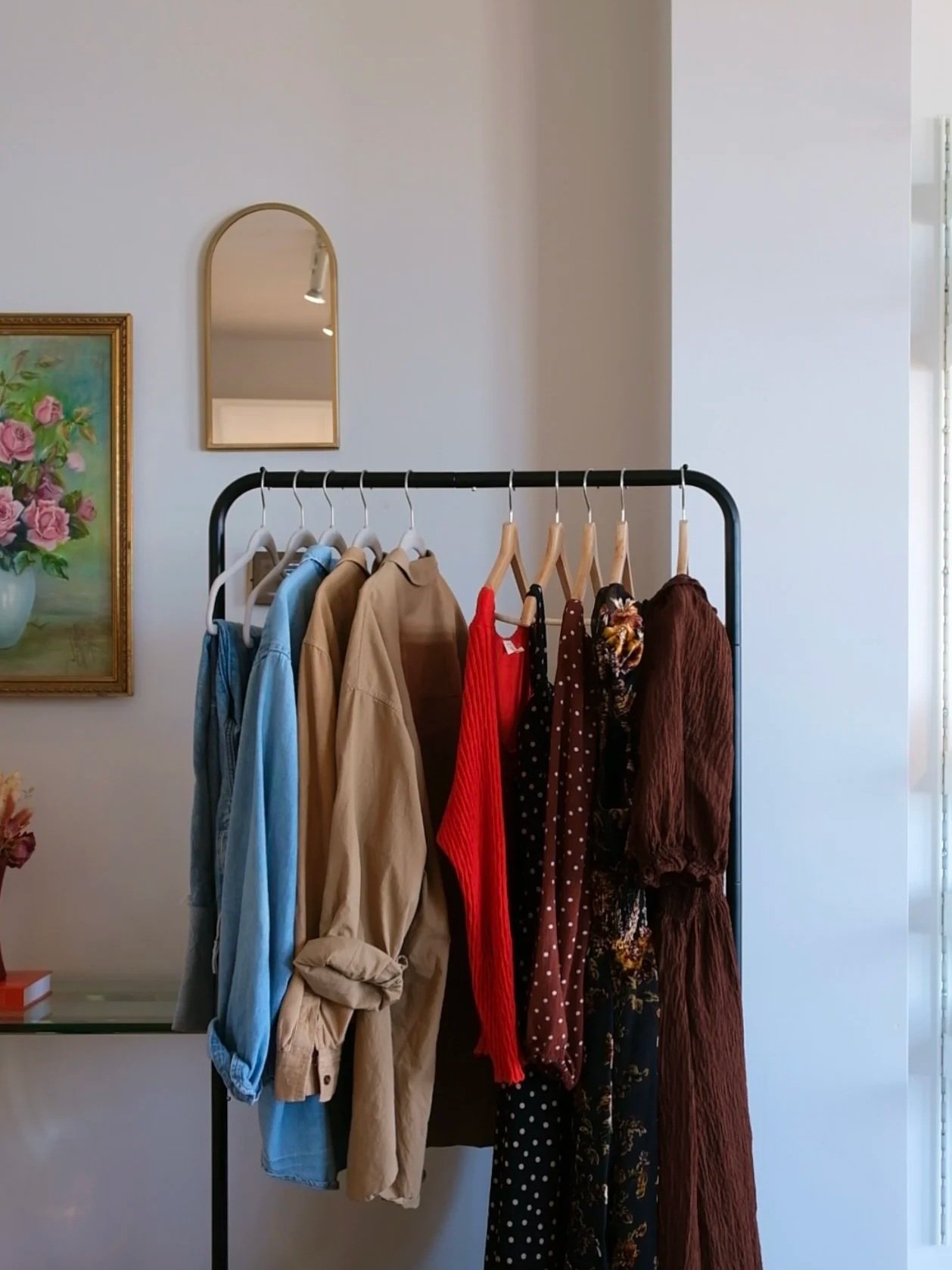 Clothing rack with various clothes, including shirts, dresses, and jackets, in a room with wall decor and a mirror.