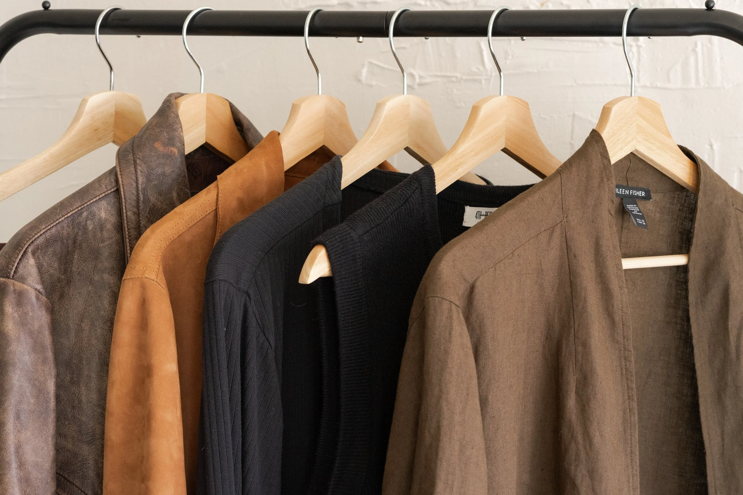 A black clothing rack holding five jackets or coats in various shades of brown, black, and gray, against a white brick wall.