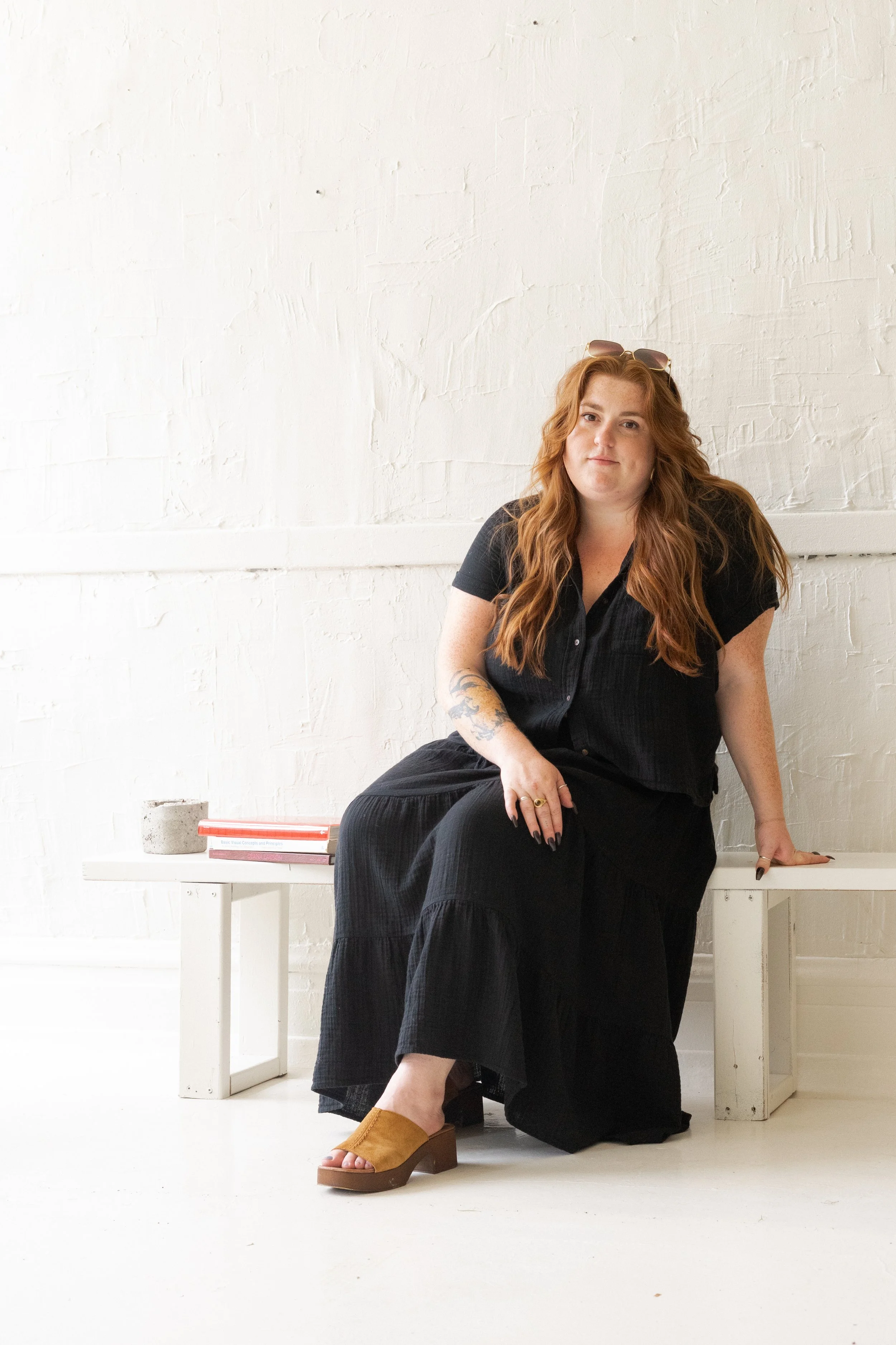 A woman with long wavy red hair, wearing sunglasses on her head, a black top, and black wide-leg pants, sits on a white bench against a white brick wall. She has tattoos on her left arm and is wearing tan platform sandals. There are a few books and a concrete weight on the floor beside the bench.