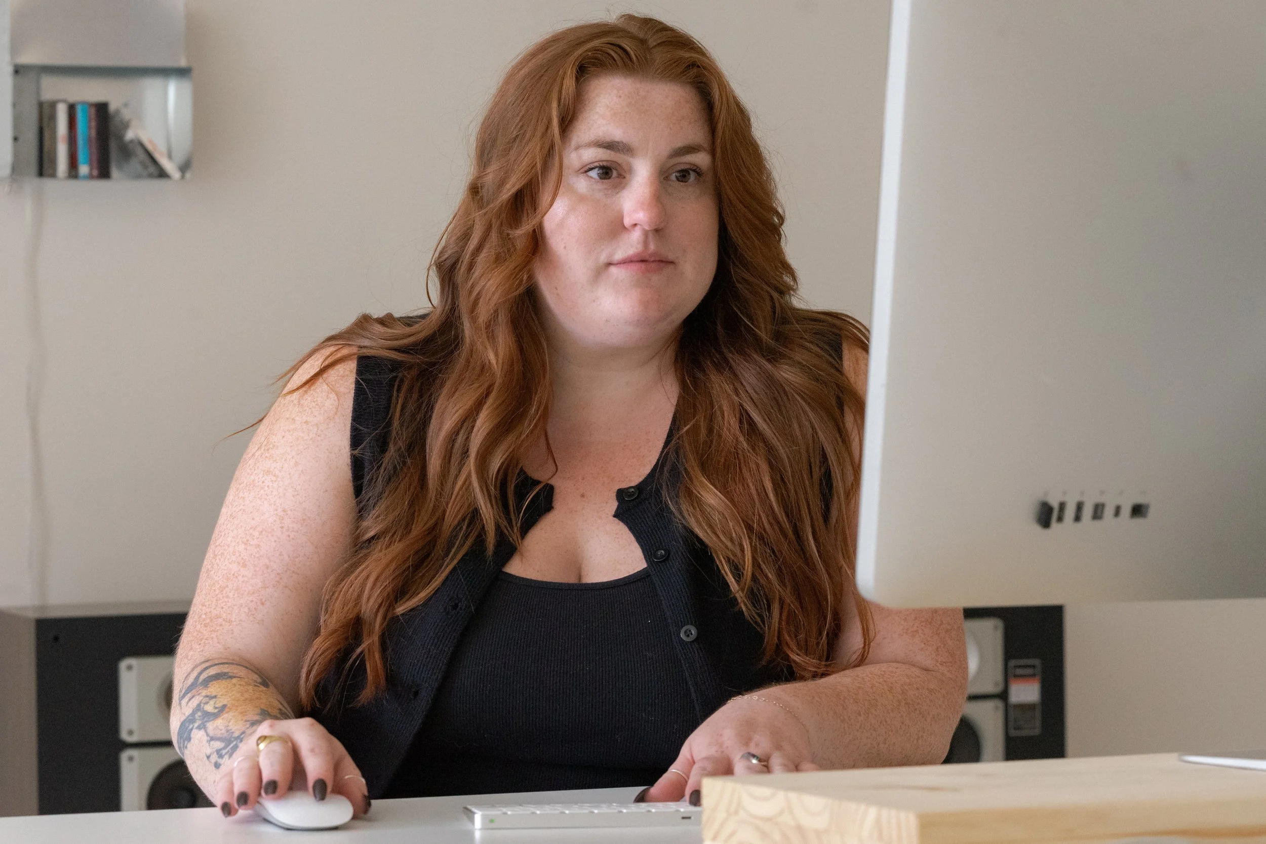 A woman with long red hair and freckles is working on a computer at a desk. She is wearing a black sleeveless top and has tattoos on her arm.