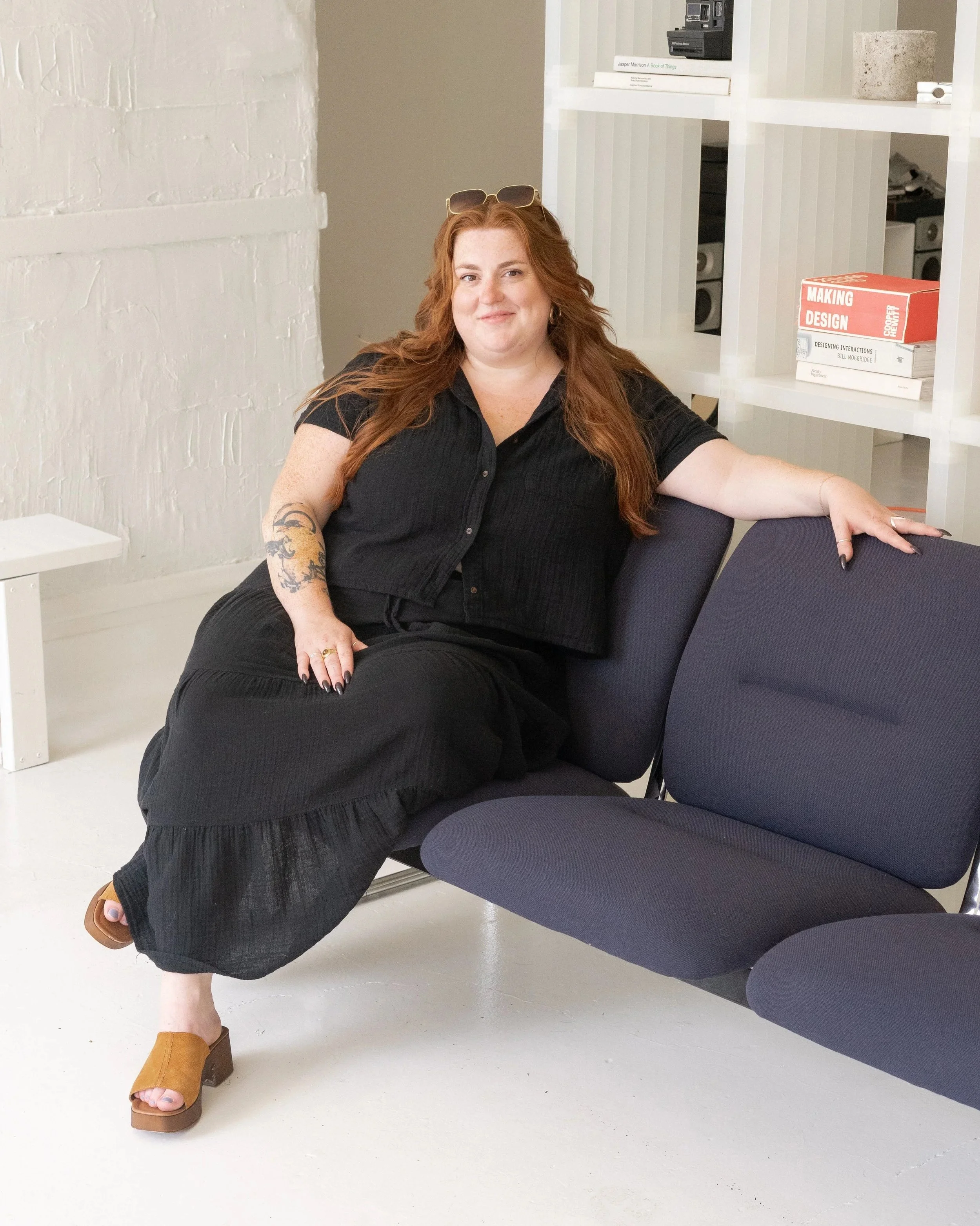 A woman with long red hair, glasses on her head, wearing a black shirt and black pants, sitting casually on a chair with one leg crossed over the other. She is smiling slightly, with jewelry on her fingers and a tattoo on her left arm. She is in a modern, minimalist room with white and beige walls, a white shelving unit with books and decor, and purple chairs.