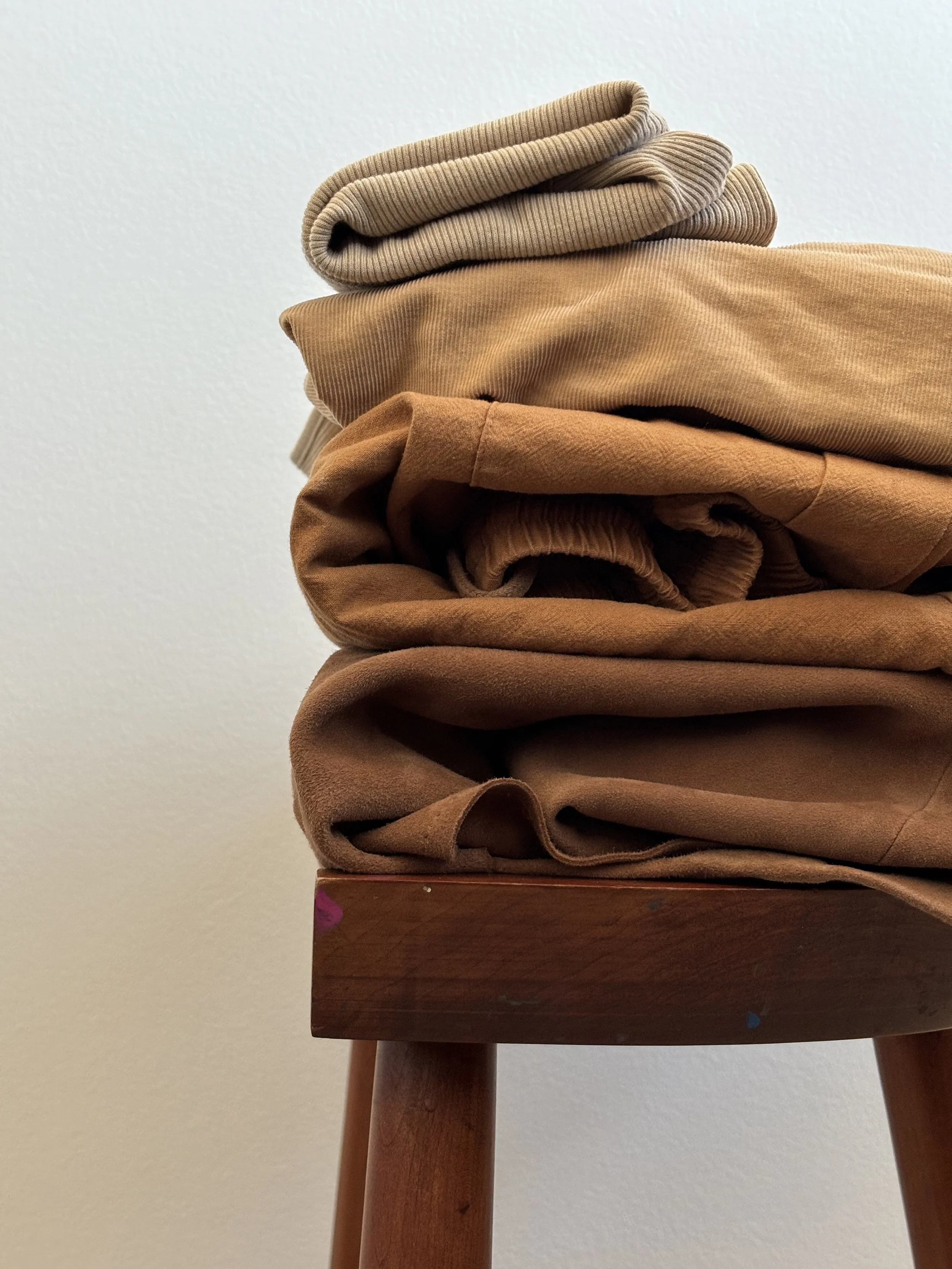 Stacked beige and brown folded clothing on a wooden table against a plain wall.