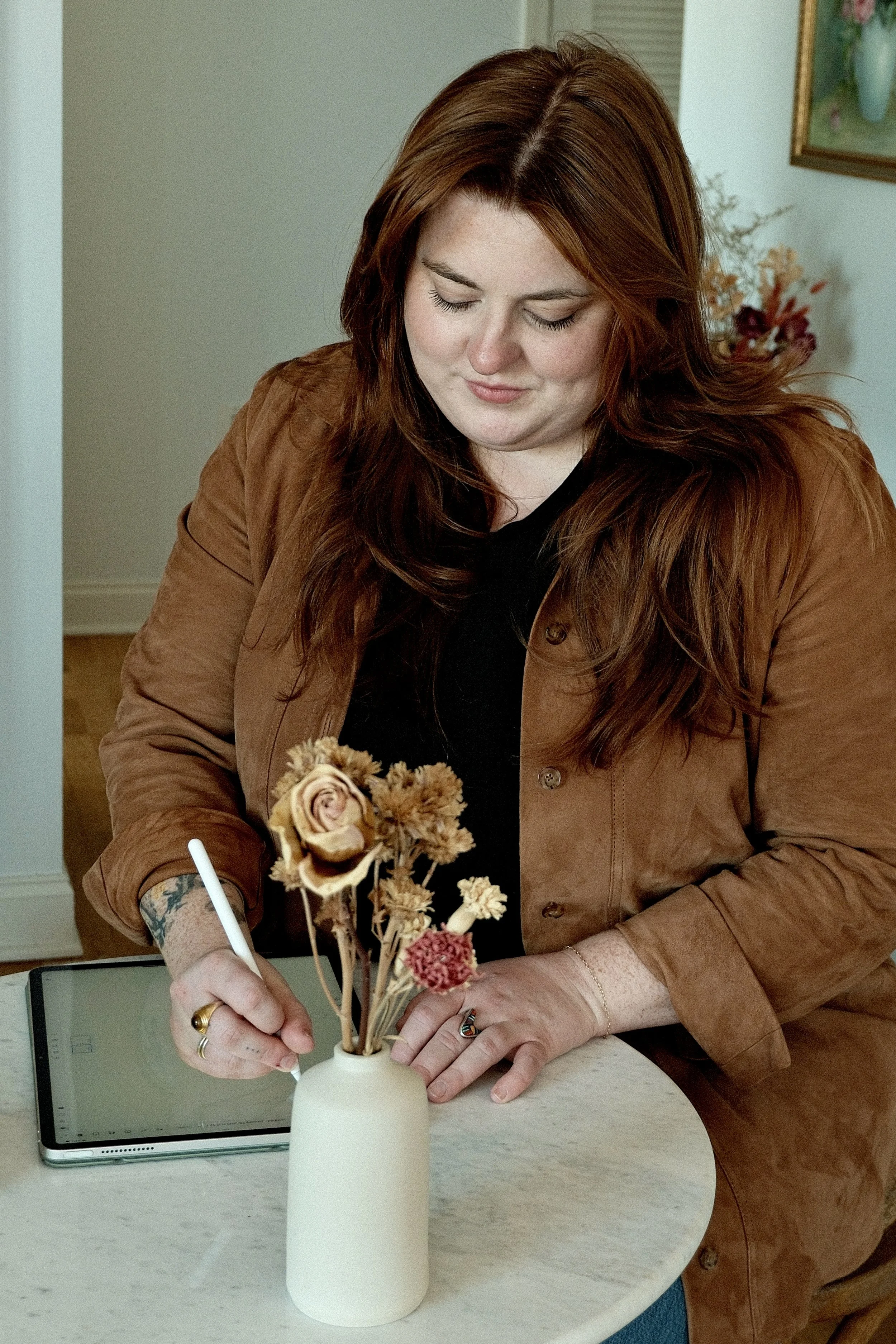 A woman with long red hair wearing a brown jacket and black shirt is sitting at a table making a floral arrangement in a white vase with dried flowers. A tablet is on the table nearby.