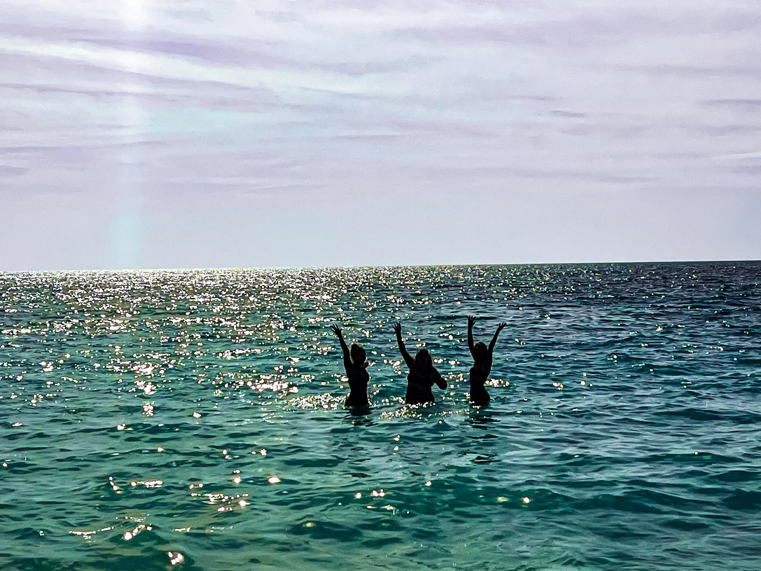 Three people in the ocean raising their hands in the air, with the sun reflecting off the water under a cloudy sky.