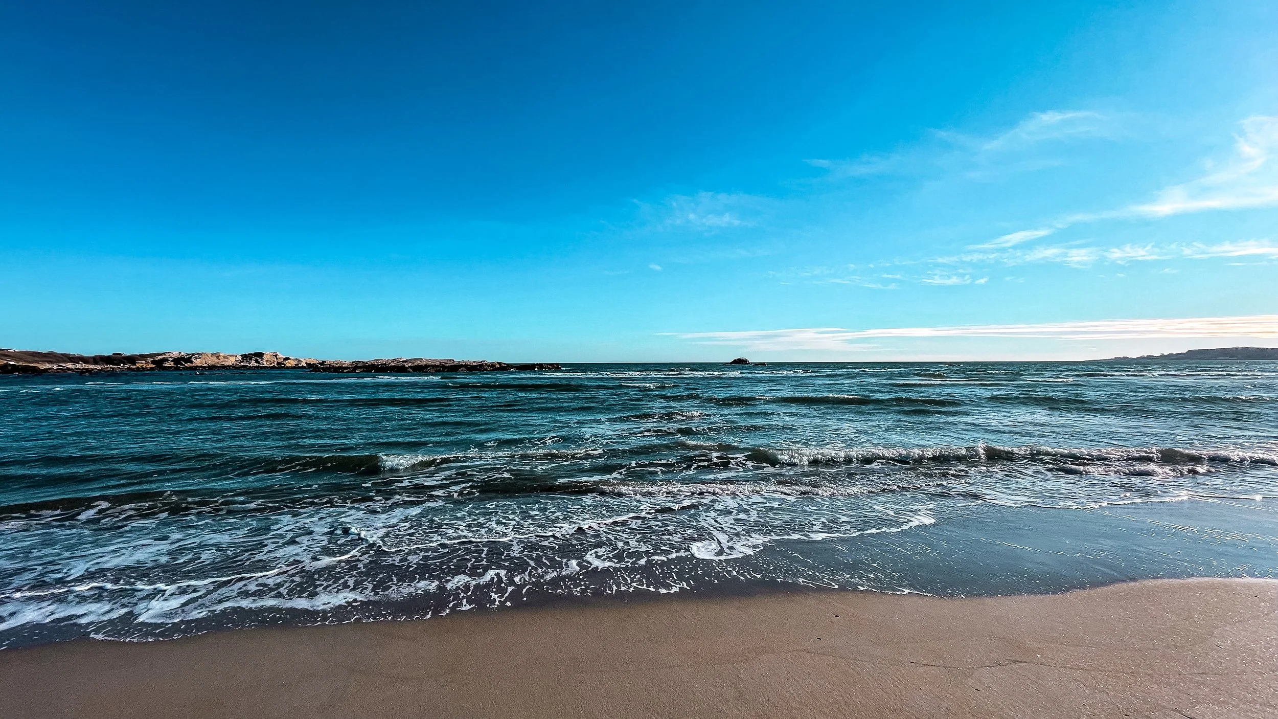 Calm ocean waves wash onto a sandy beach under a clear blue sky with wispy clouds.