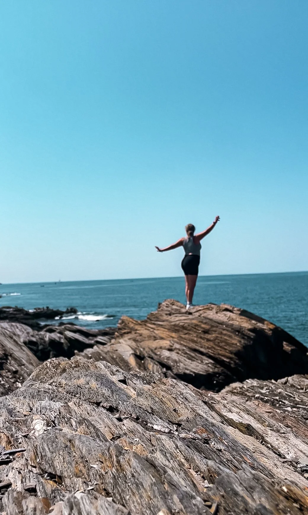 Woman in black shorts and a sleeveless top standing on a rocky shoreline with arms outstretched, facing the ocean under a clear blue sky.
