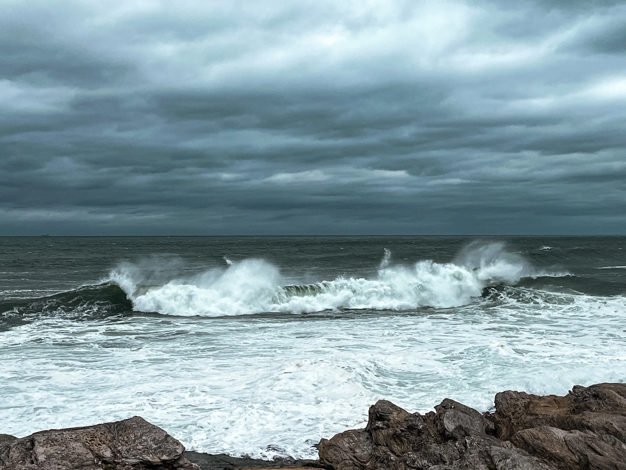 Overcast sky above choppy ocean waves crashing against rocks on the shoreline.