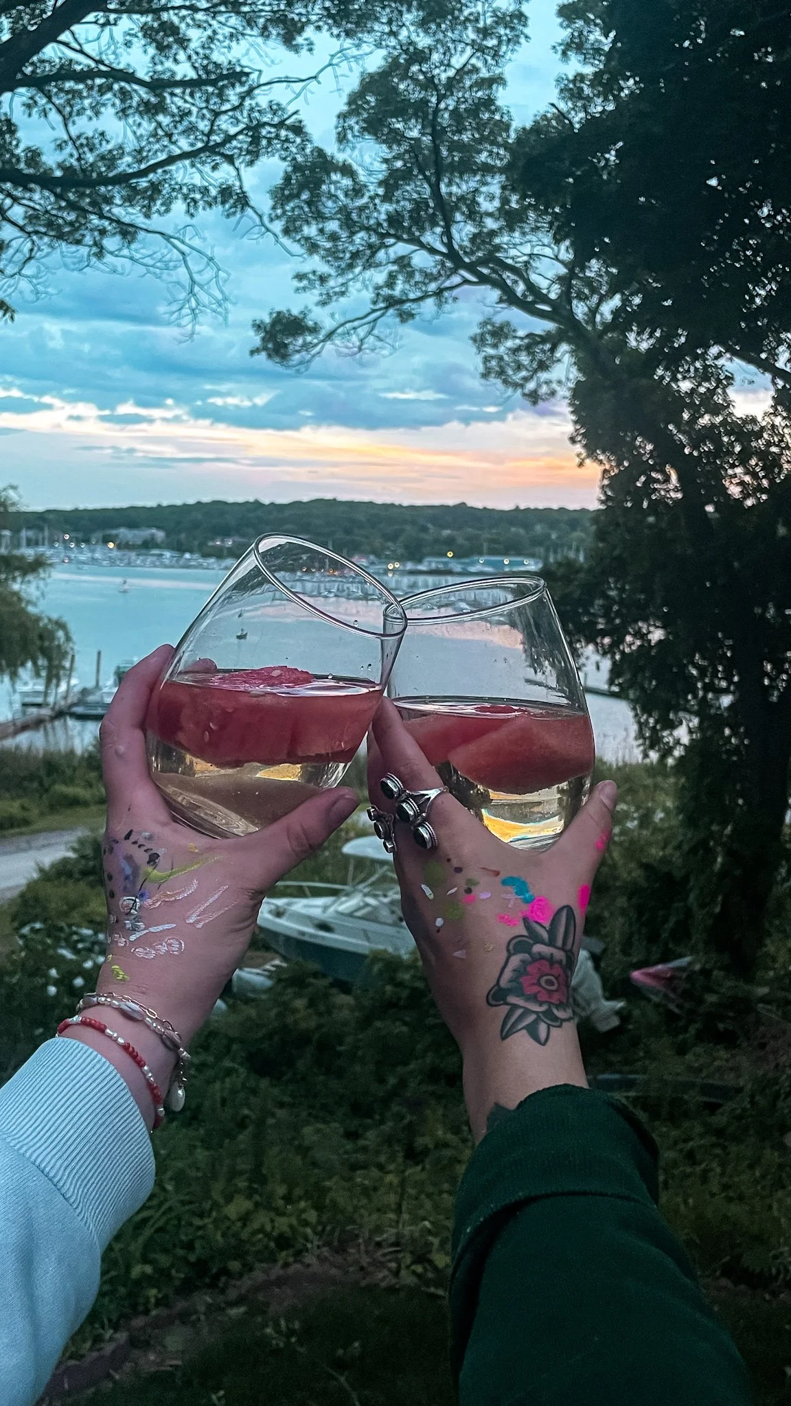 Two hands holding glasses of white wine or champagne with peach slices, raised in a toast outdoors at sunset, with trees, water, and boats in the background.