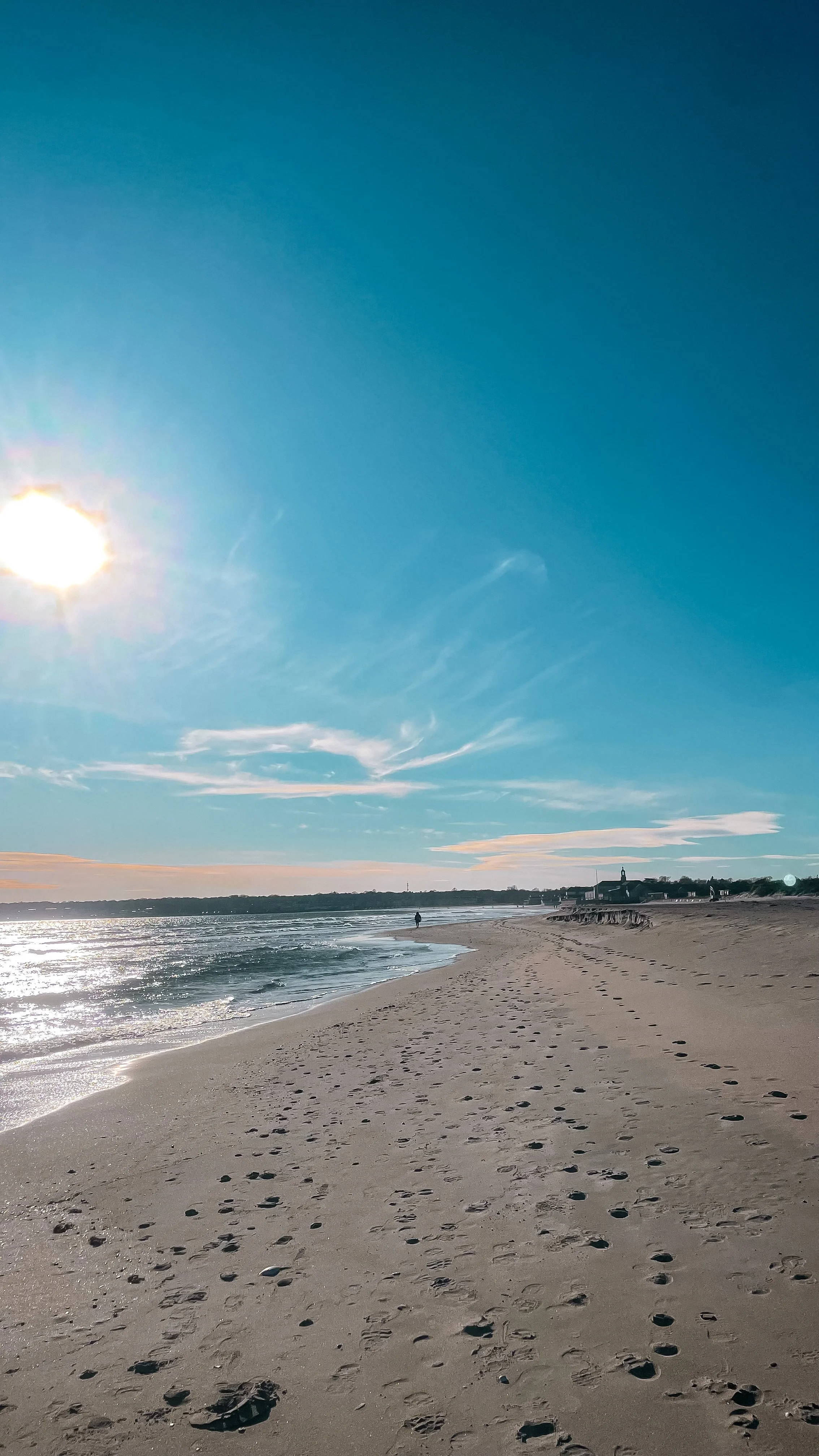 A beach scene with footprints in the sand, gentle waves, a person walking along the shoreline, and buildings in the distance under a clear blue sky with some clouds and the sun shining.
