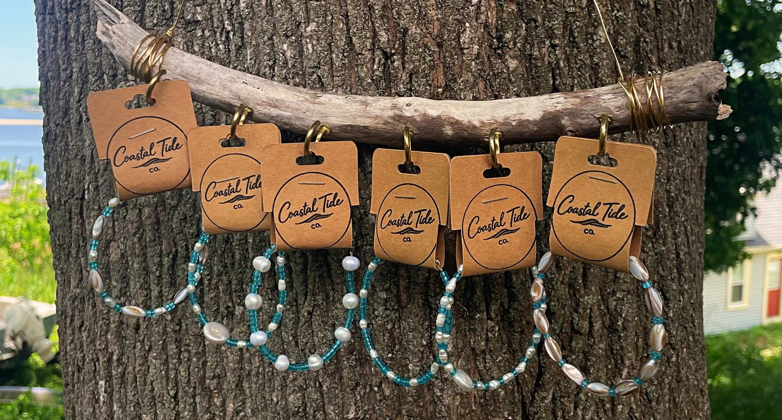 Jewelry necklaces hanging on a log attached to a tree trunk, with tags saying 'Coastal Tide Co.'