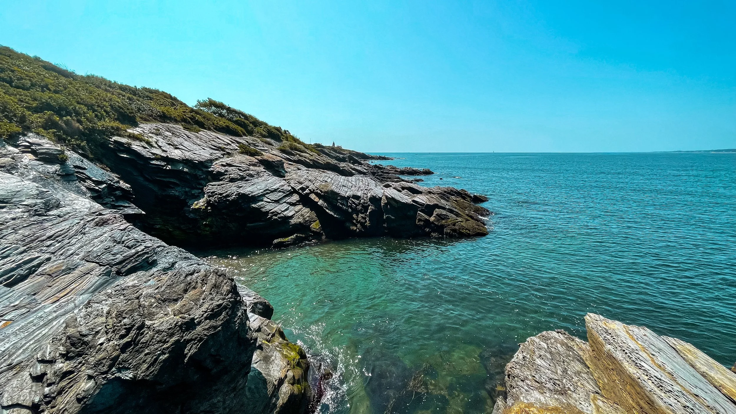 Rocky shoreline with clear water and a distant horizon under a bright blue sky.