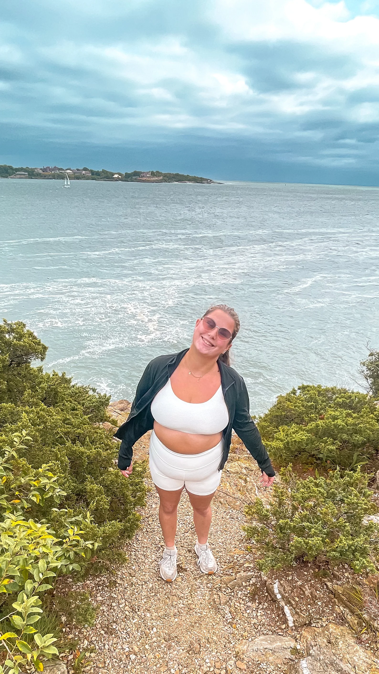 A woman standing on a rocky trail near the water, smiling, with bushes in the foreground and a large body of water with a sailboat and a distant shoreline in the background.