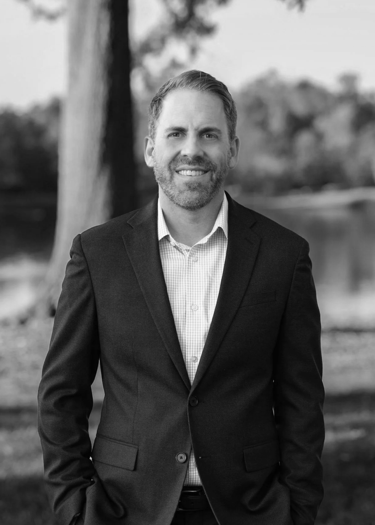 A black and white portrait of a man with a beard, wearing a suit jacket and a checked shirt, standing outdoors with trees and a body of water in the background.