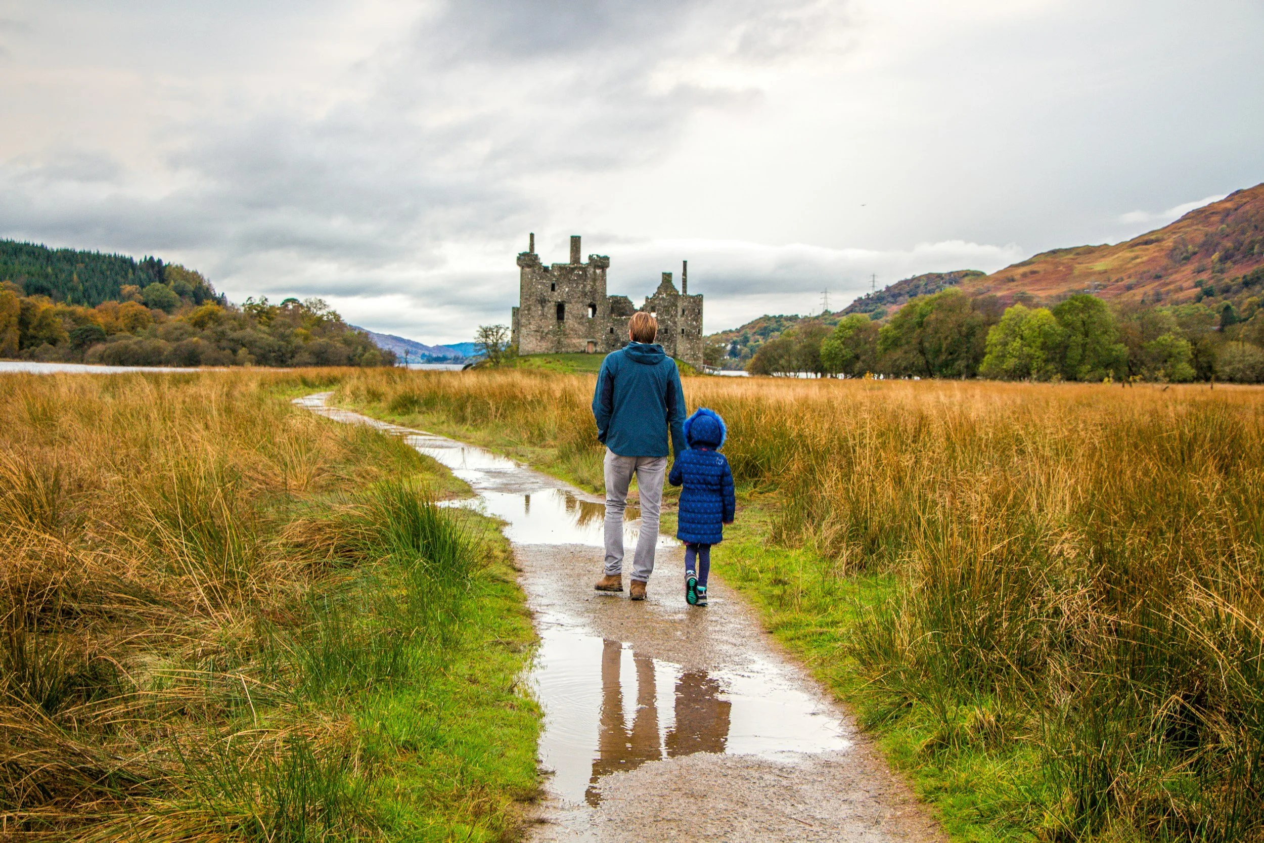 A man and a child walking on a muddy path surrounded by grass, heading towards a castle in a hilly, autumn landscape with cloudy skies.