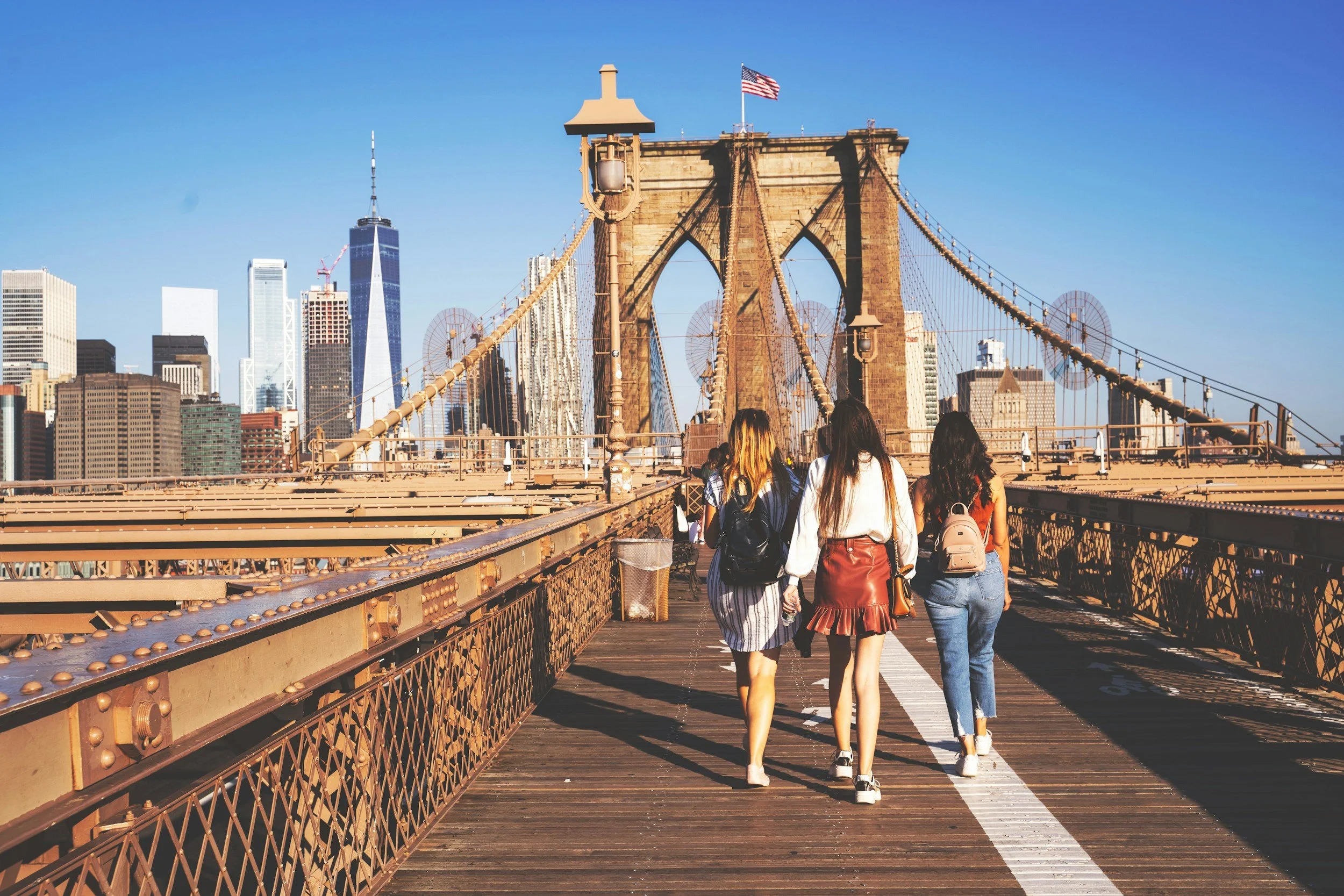 Four women walk across the Brooklyn Bridge in New York City with city skyscrapers, including One World Trade Center, in the background.
