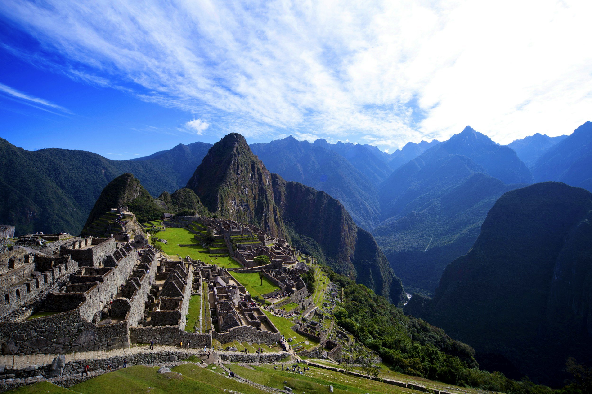 Ancient Incan ruins at Machu Picchu surrounded by green mountain ridges and a bright blue sky with wispy clouds.