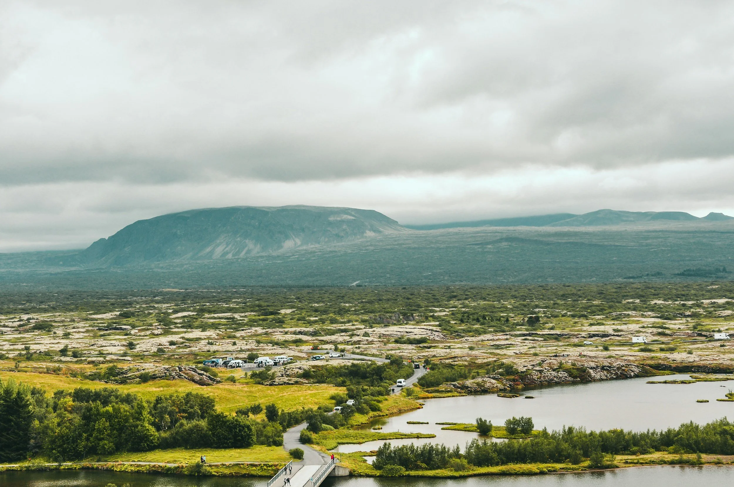 Landscape featuring a mountain with a flat top, cloudy sky, a river, green fields, and a small parking lot with vehicles.
