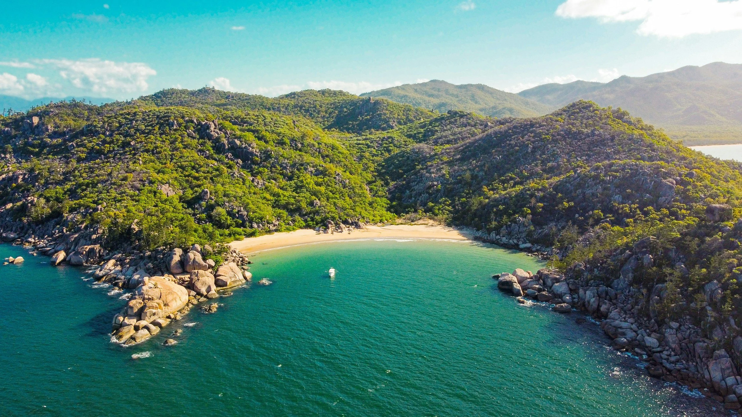 Aerial view of a tropical coastline with a sandy beach surrounded by rocky hills covered in dense greenery, clear blue water, and distant mountains under a partly cloudy sky.