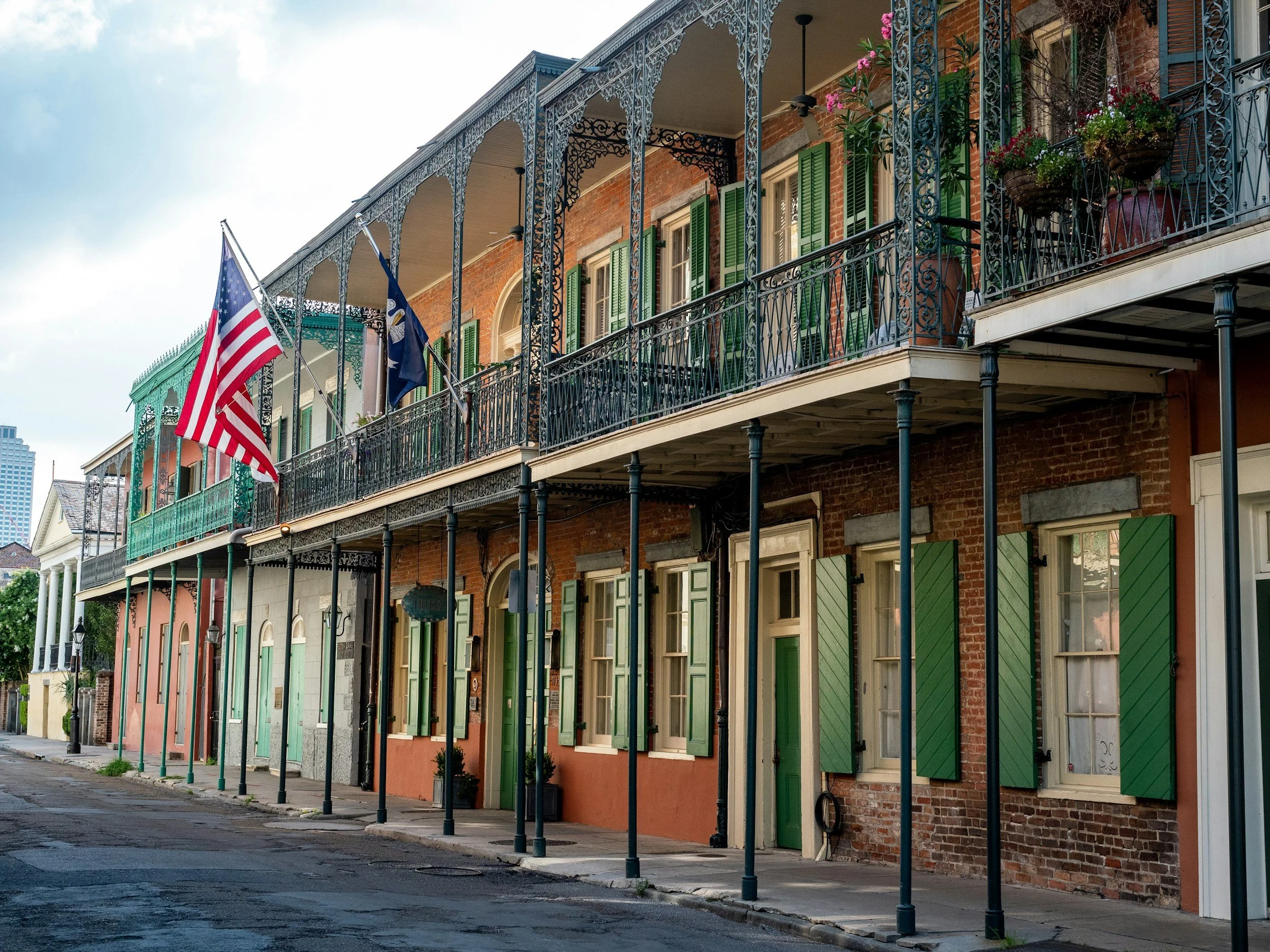 A row of historic brick buildings with colorful green, pink, and cream shutters on the windows and decorative wrought iron balconies. Two American flags are hanging from the balconies. The street in front is empty with a cloudy sky overhead.