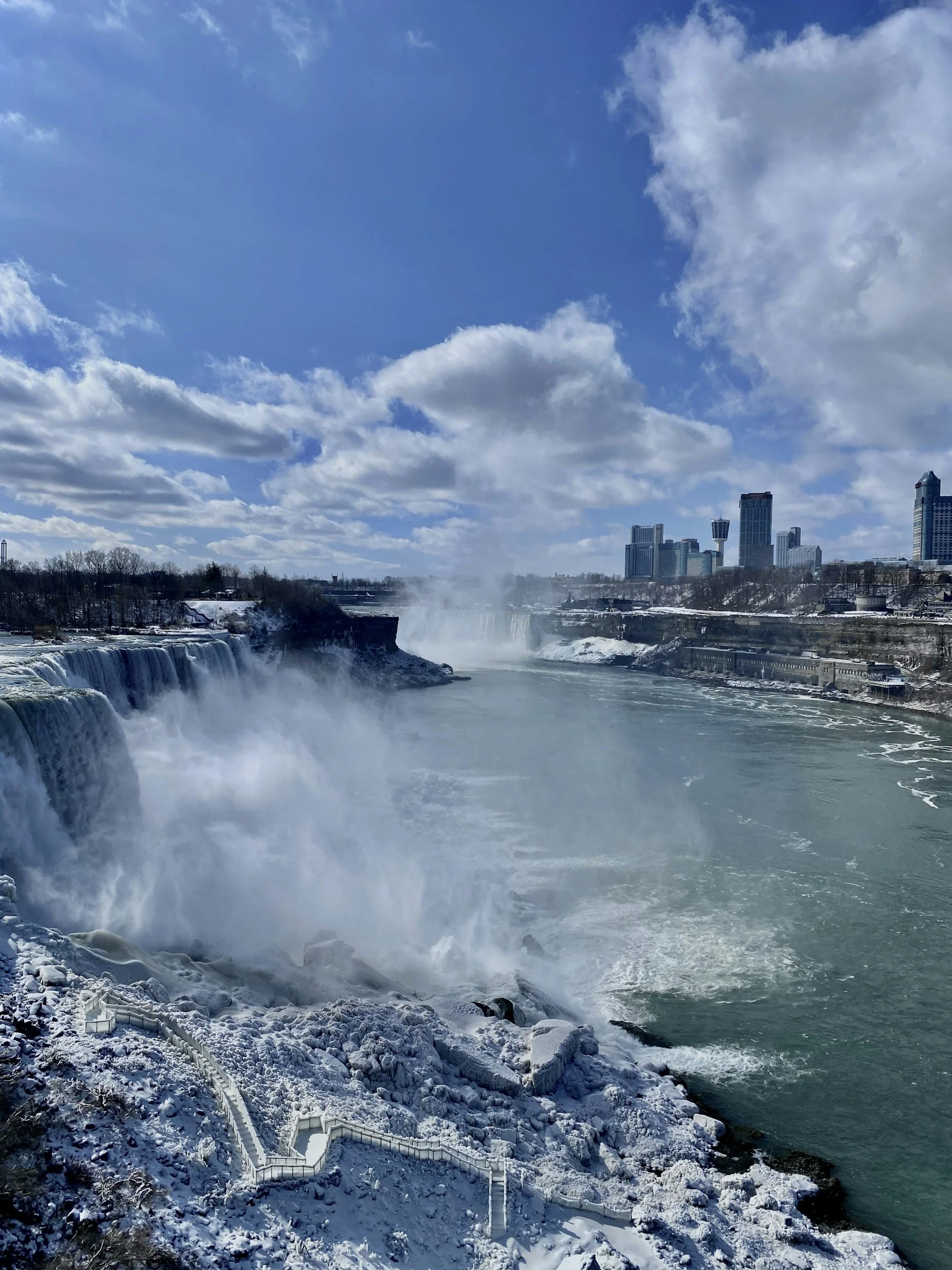 Snow-covered landscape with waterfalls, city skyline, and snowy paths, under a partly cloudy sky.