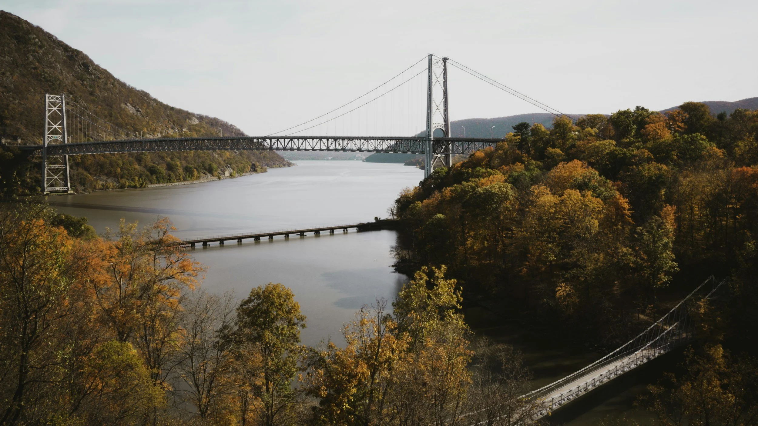 A large suspension bridge over a river, surrounded by colorful autumn trees and hills in the background.