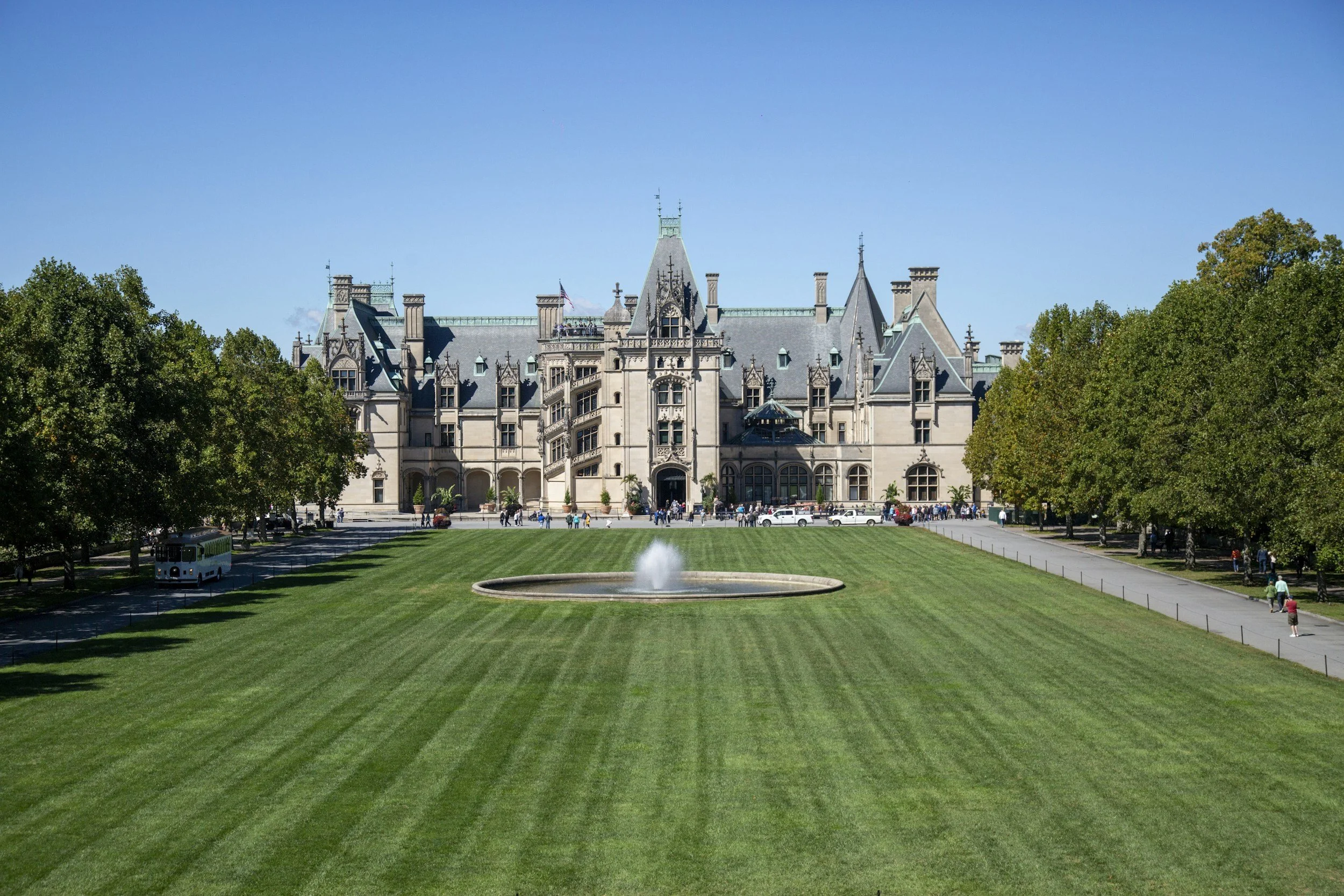 A large, historic castle-like mansion with multiple turrets, set behind a well-manicured lawn with evenly cut grass, a fountain in the foreground, and trees lining the sides. The sky is clear and blue.