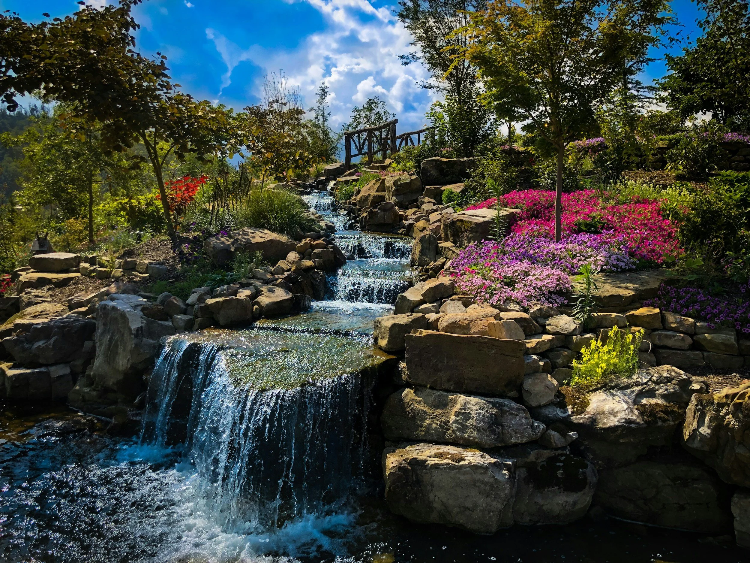 A vibrant garden with a small cascading waterfall flowing over rocks, surrounded by colorful flowers and green trees under a bright blue sky with fluffy white clouds.