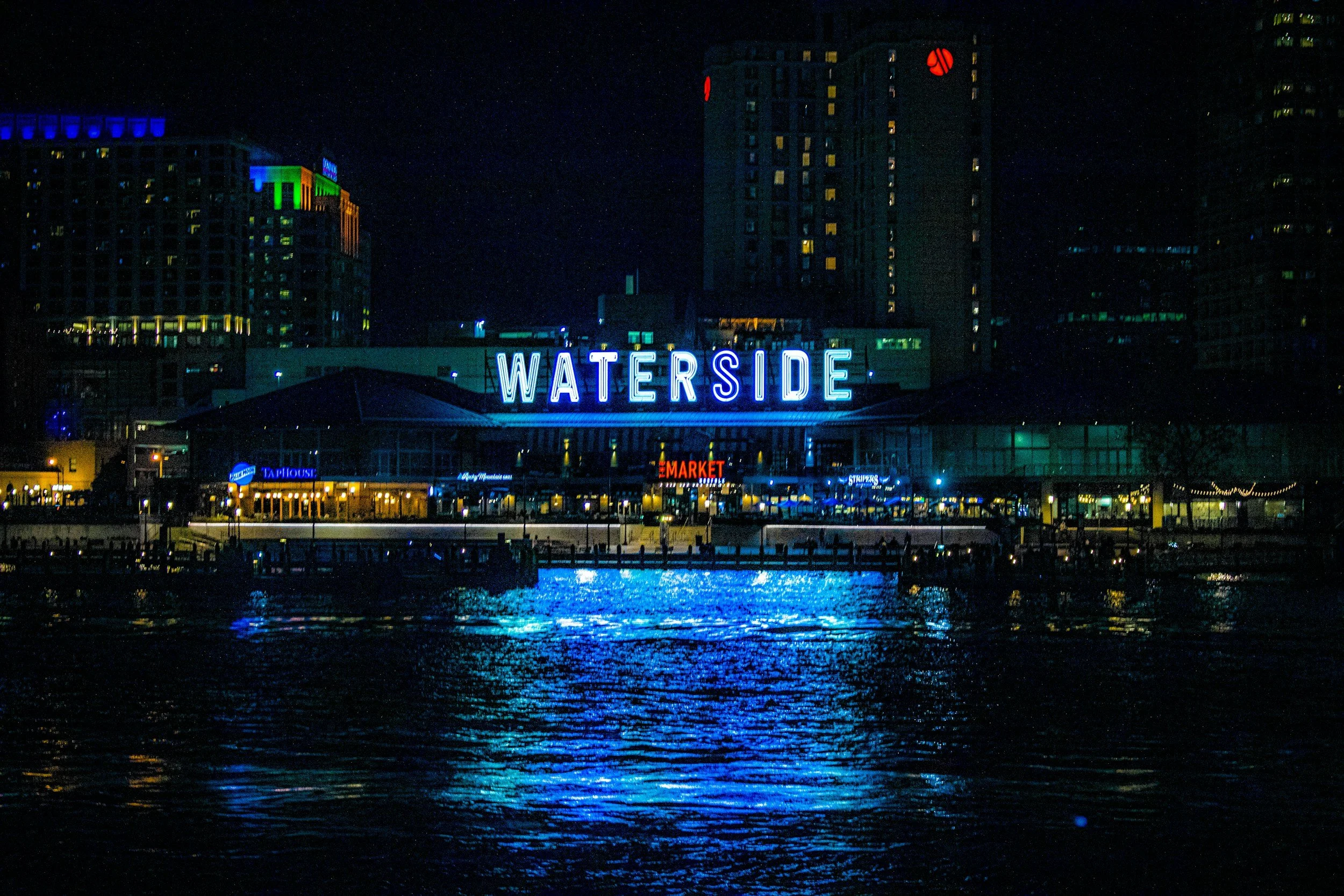 Night view of a city waterfront with illuminated buildings, including a prominent neon sign that says 'WATER SIDE', reflecting in the water below.