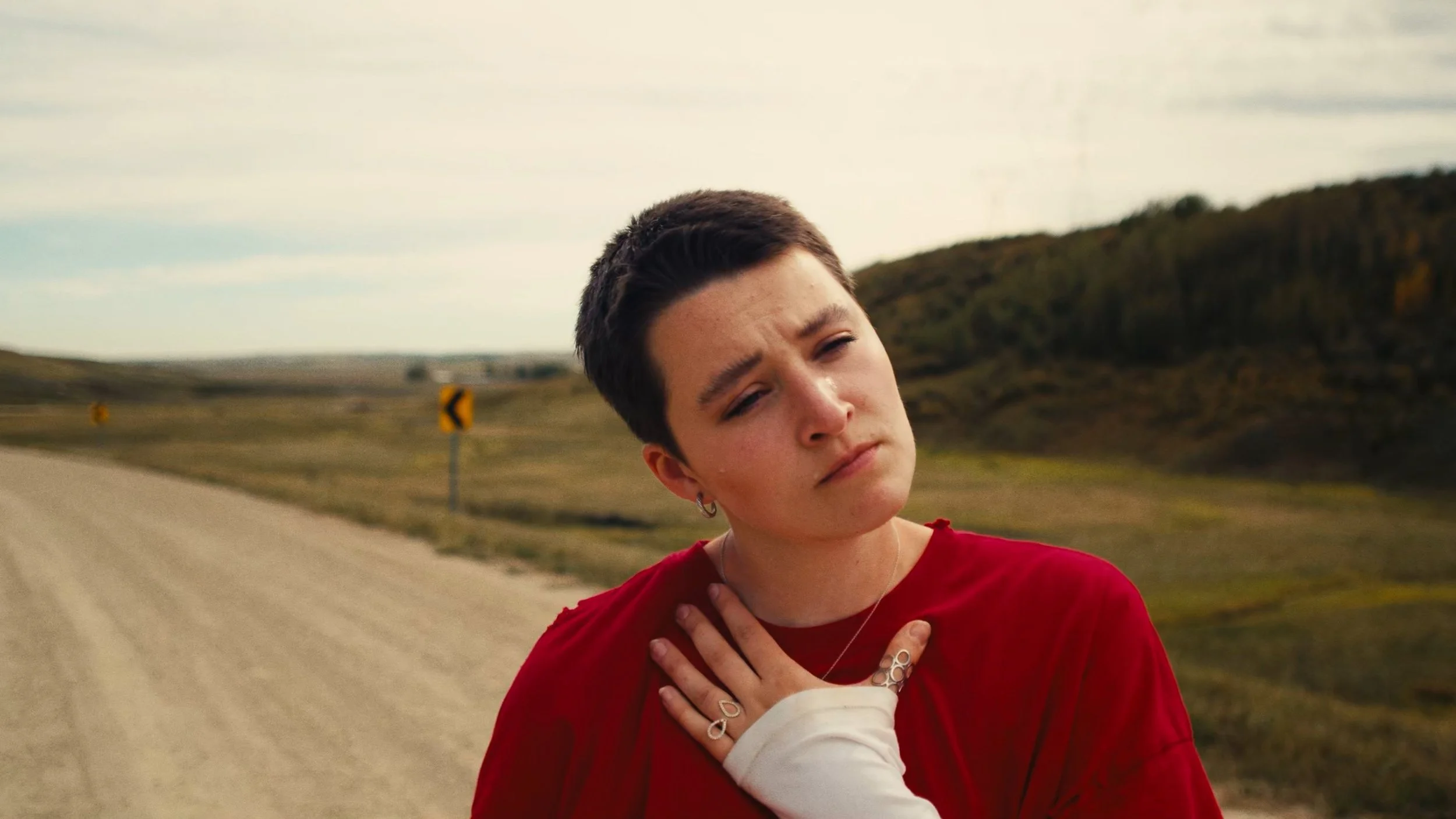 A young person with short dark hair and earrings wearing a red shirt is standing outdoors on a rural road with a backdrop of grassy hills and cloudy sky.