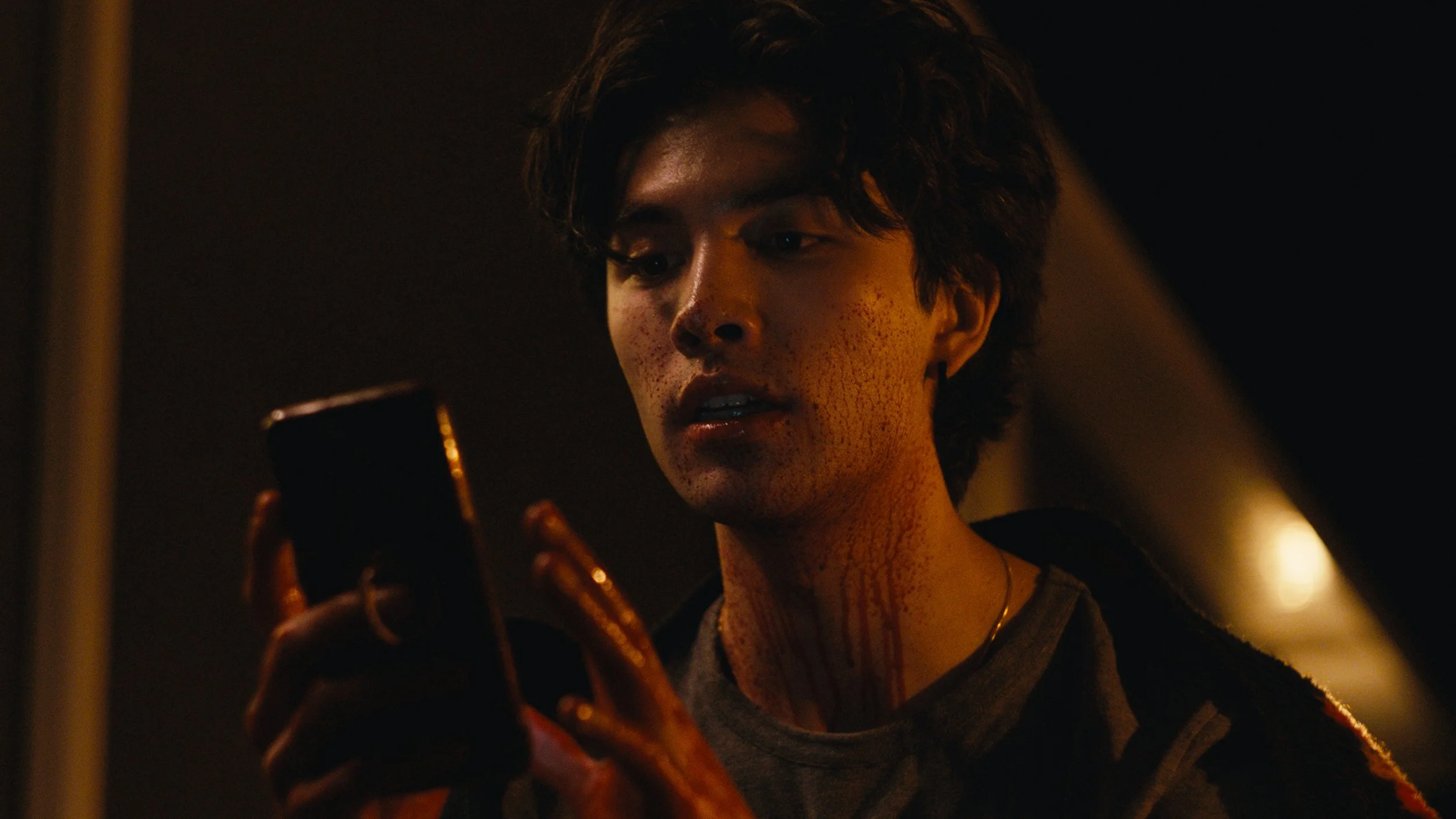 A young man with dark, wavy hair and a distressed expression, looking at his phone in a dimly lit environment.