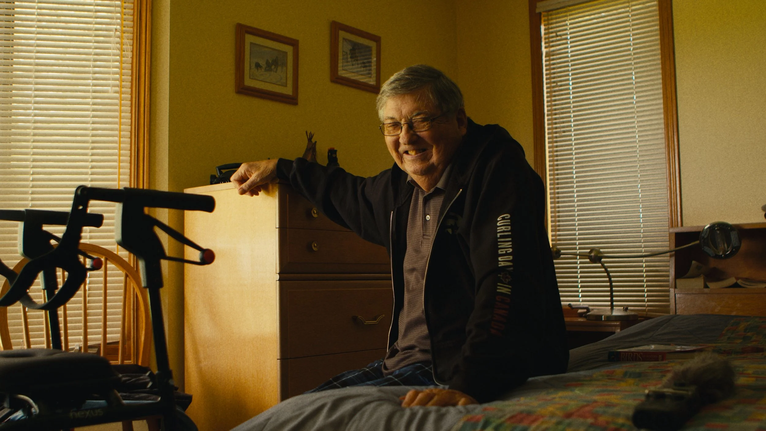 An elderly man with glasses smiling and reclining on a bed in a warmly lit bedroom, with wooden blinds and framed pictures on the wall.