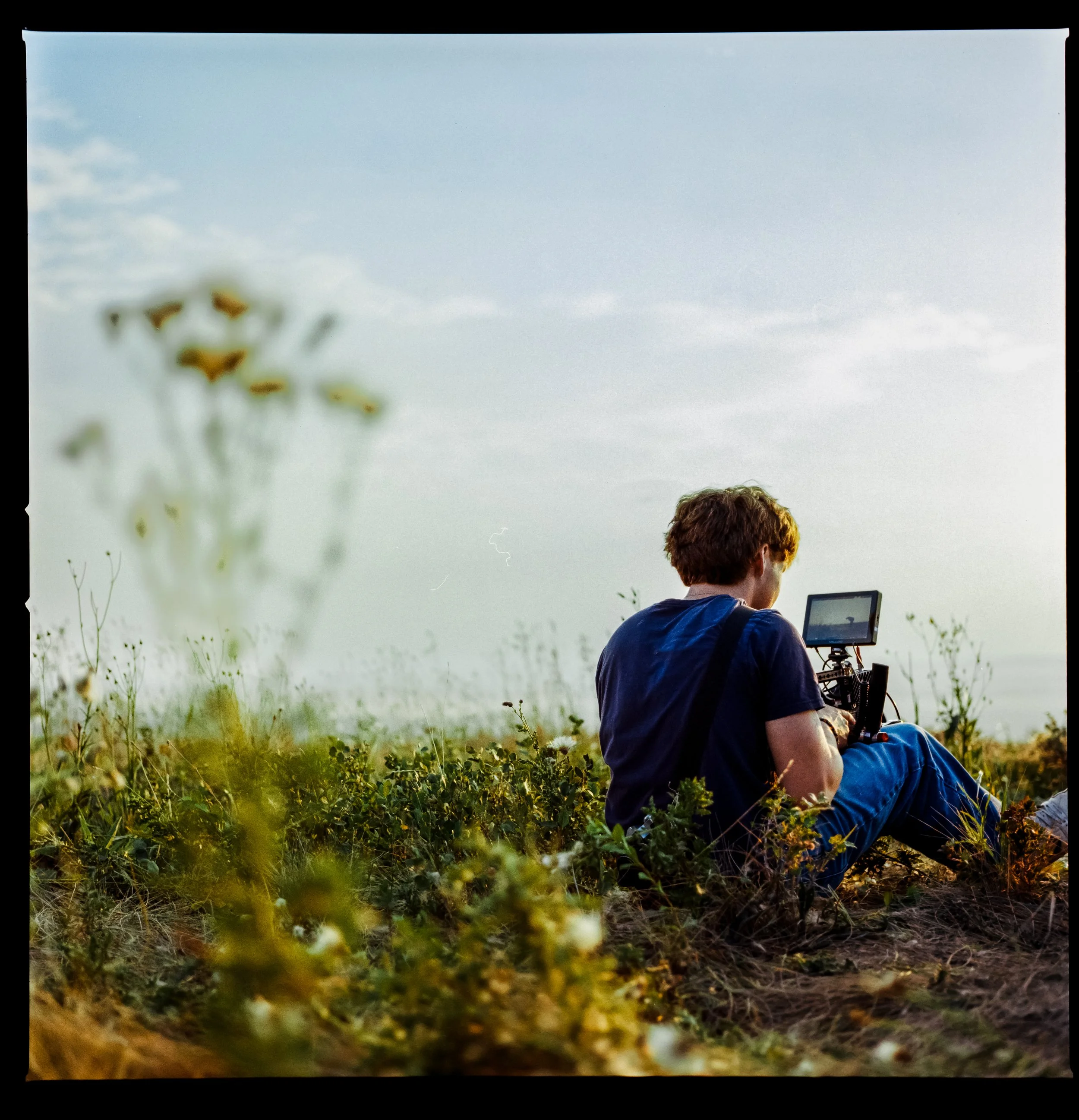 A young man sitting on the ground outdoors, focused on a tablet, with tall grass and plants in the foreground and a blue sky with some clouds in the background.