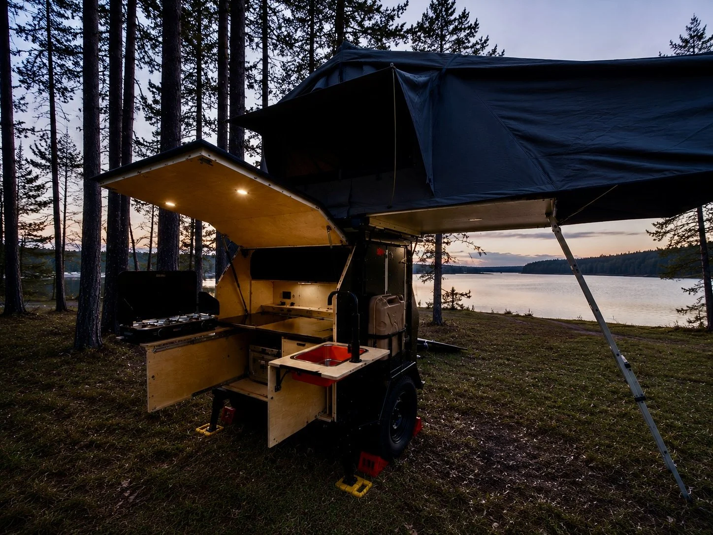 A mobile outdoor kitchen unit with a black cover set up in a wooded area near a lake at sunset.