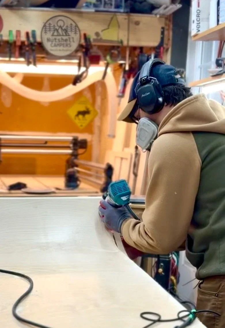 Person wearing a cap, headphones, and a face mask, working with a power tool on a light-colored work surface inside a woodworking shop.