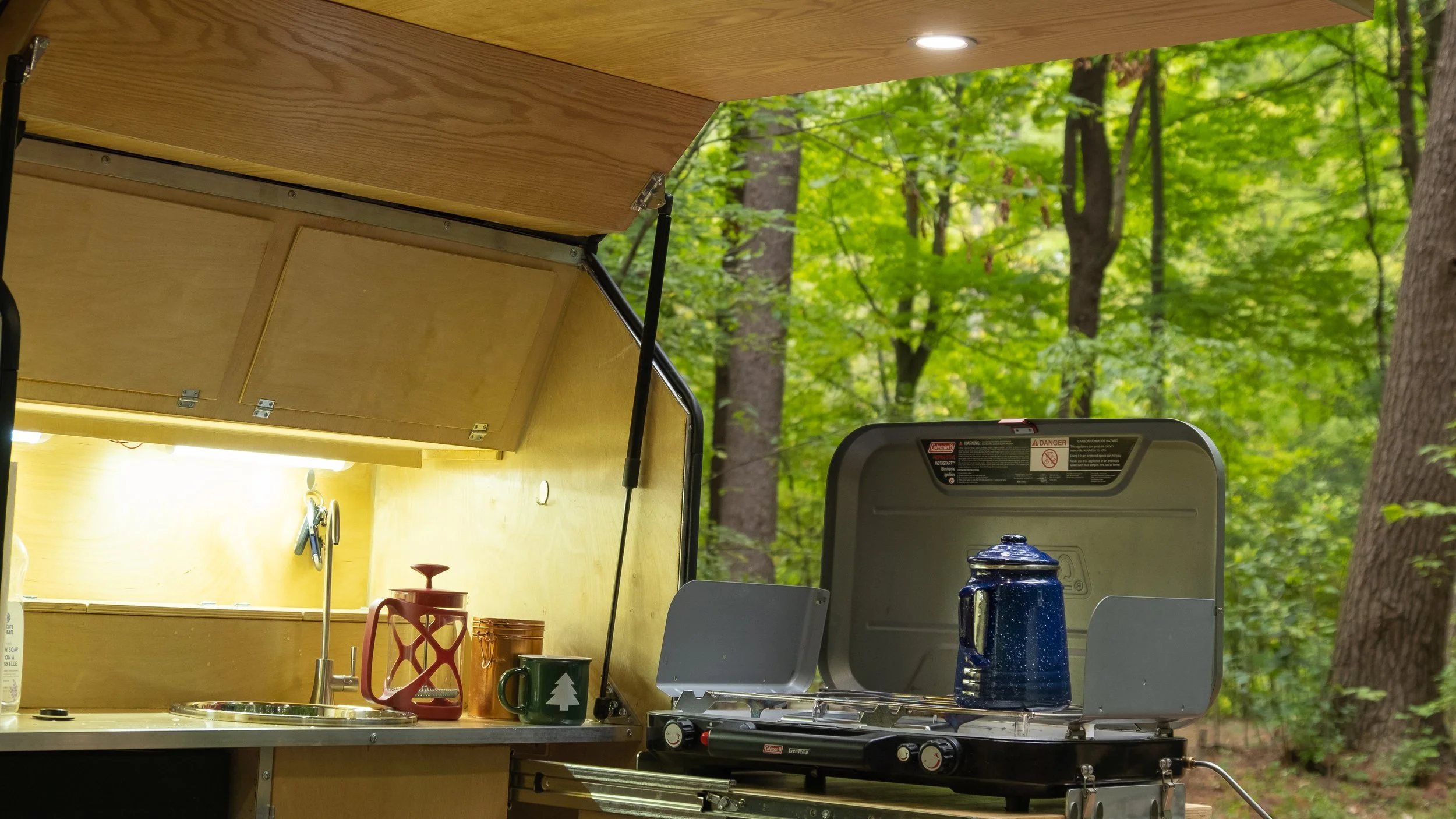 Open wooden camping stove with kitchen items on a countertop, set in a forest with green trees in the background.