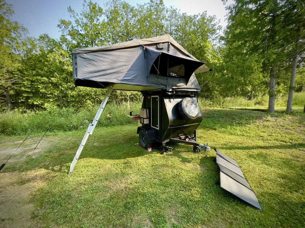 A compact travel trailer with a rooftop tent set up on a grassy area, surrounded by trees.