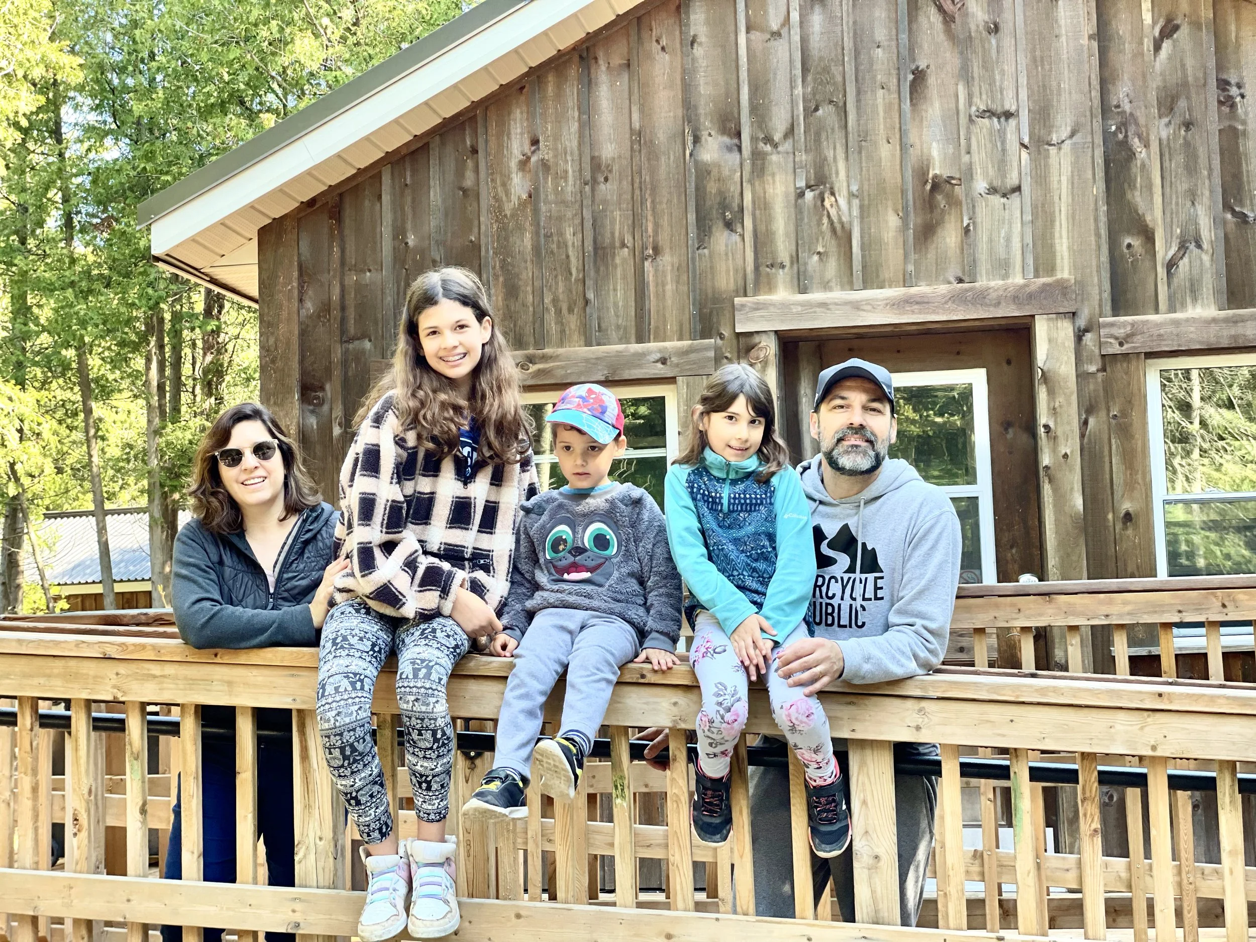 Group of seven people, including children and adults, sitting on a wooden deck in front of a wooden house with a sloped roof, surrounded by trees with green leaves, smiling at the camera.