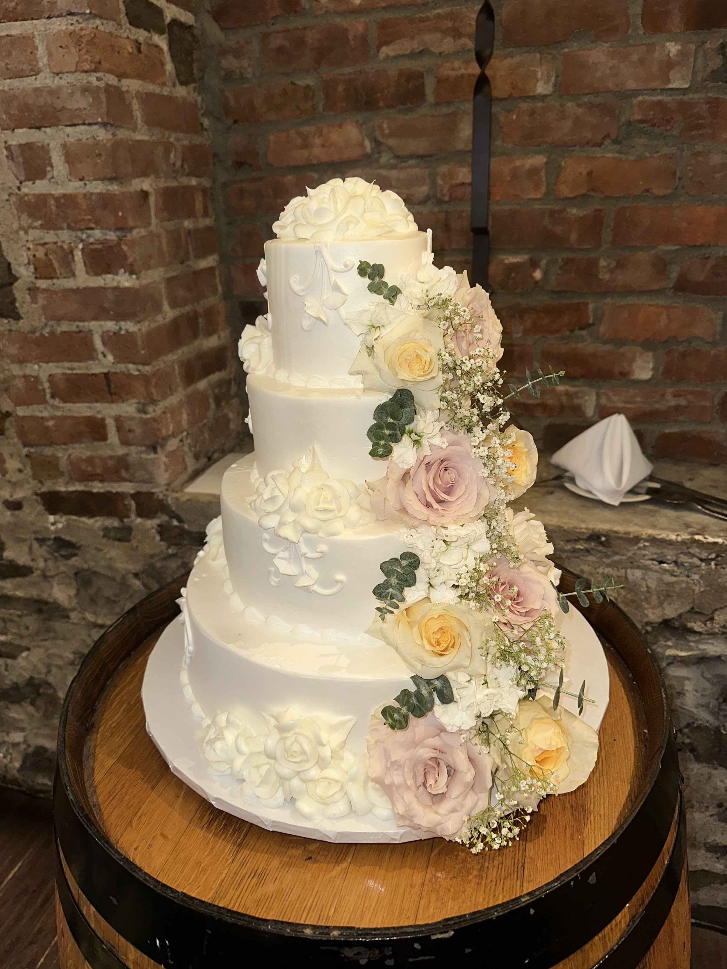 A three-tier white wedding cake decorated with cream-colored roses, pink roses, and green foliage, set on a wooden table against a brick wall.