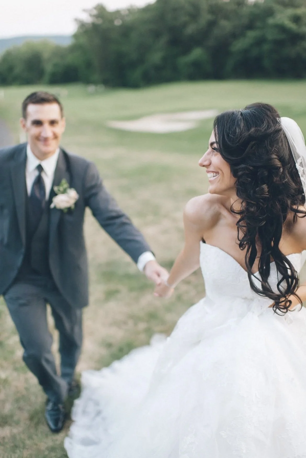 A smiling bride and groom holding hands and running outdoors on a grassy field, with trees in the background, during a wedding celebration.