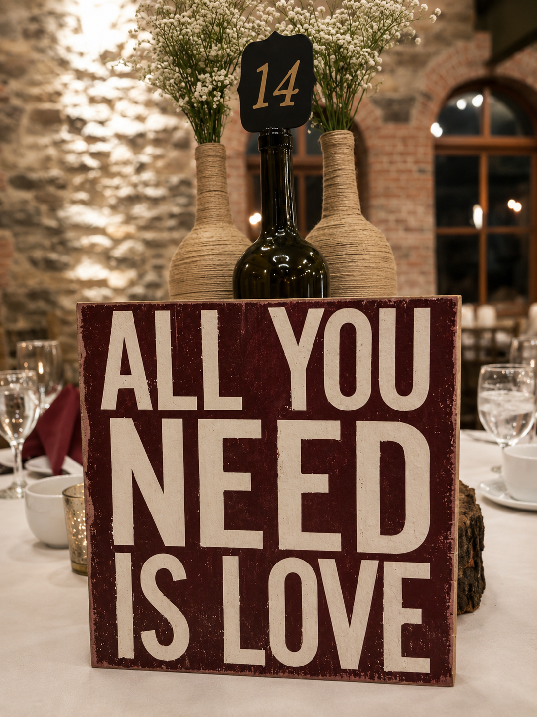 A sign on a table that reads 'ALL YOU NEED IS LOVE' with flowers and vases in the background, set in a rustic restaurant or event venue.