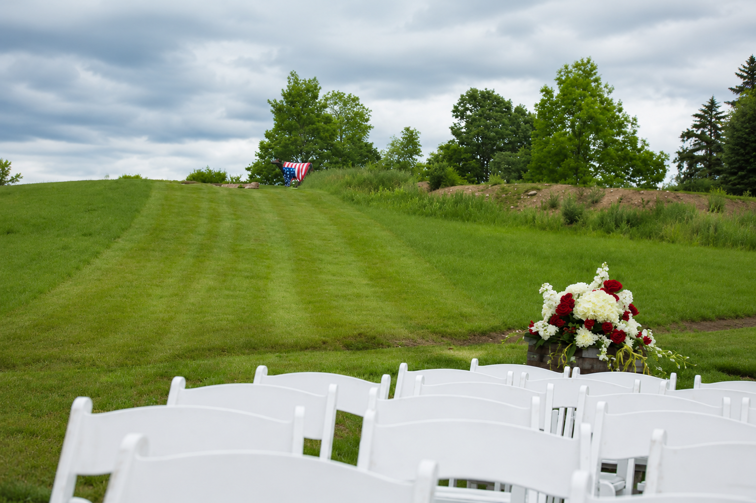 An empty outdoor wedding ceremony setup with white chairs, a flower arrangement with red and white roses, and a grassy hill with an American flag at the top, under a cloudy sky.