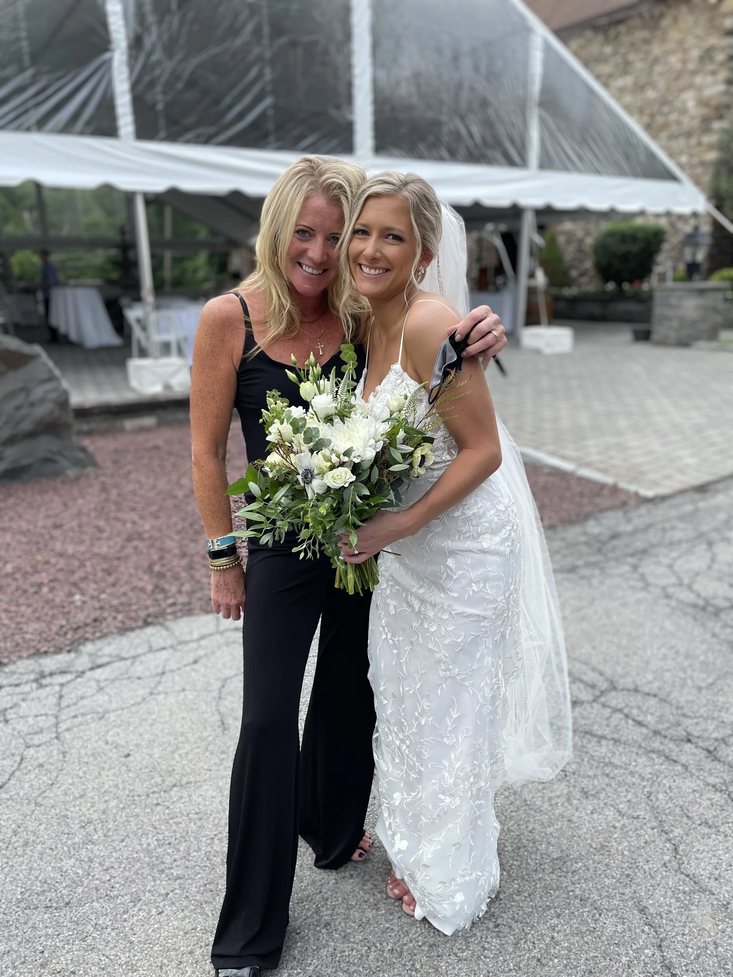 Two women, one in a black dress and the other in a wedding dress, stand close together smiling for the camera. The woman in the wedding dress holds a bouquet of white and green flowers. They are outdoors near a pavilion with a stone wall in the background.
