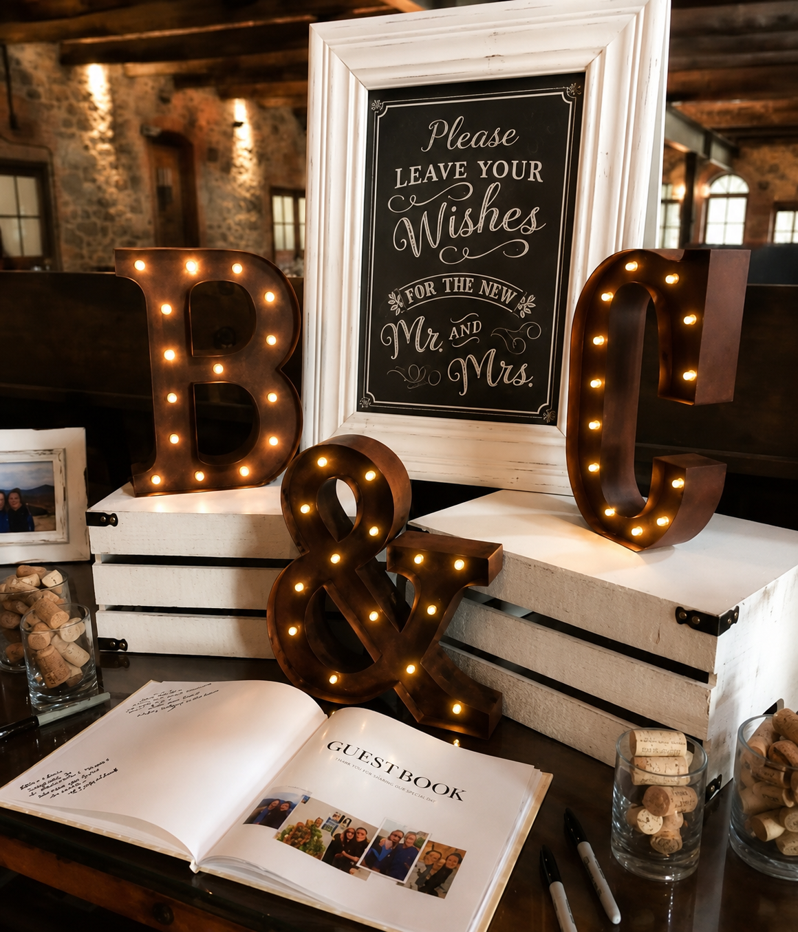 Decorative wedding display with illuminated letters spelling B, &, C, a guest book, framed photographs, and a sign that reads 'Please leave your wishes for the new Mr. and Mrs.' in a rustic venue with stone walls and wooden beams.