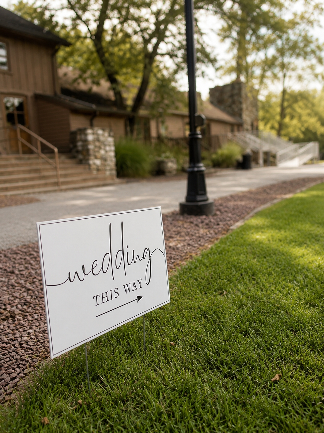 A white sign with black writing that says "wedding this way" with an arrow pointing to the right, placed on green grass outside, with a gravel pathway, lamppost, and brown wooden houses in the background.