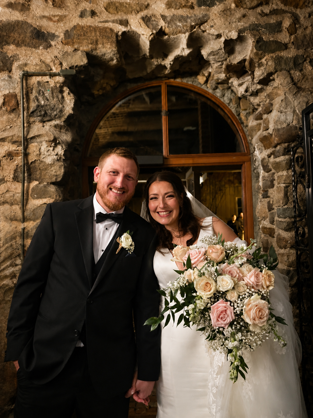 A happy newlywed couple stands in front of a stone wall, with the bride holding a large bouquet of pink and white roses and the groom dressed in a black tuxedo.